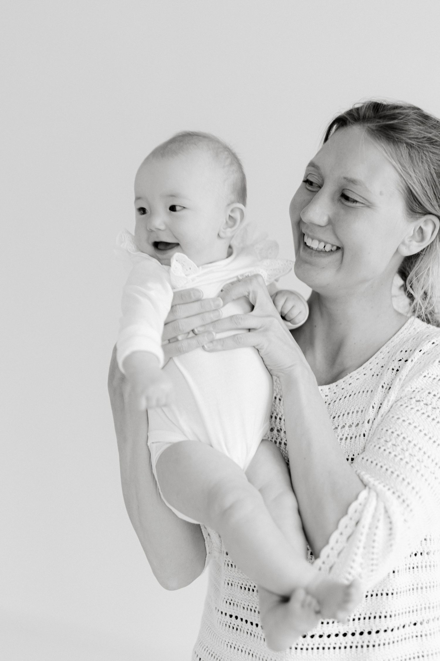 Black and white photo of a smiling mother holding her happy baby in a white onesie against a light background.
