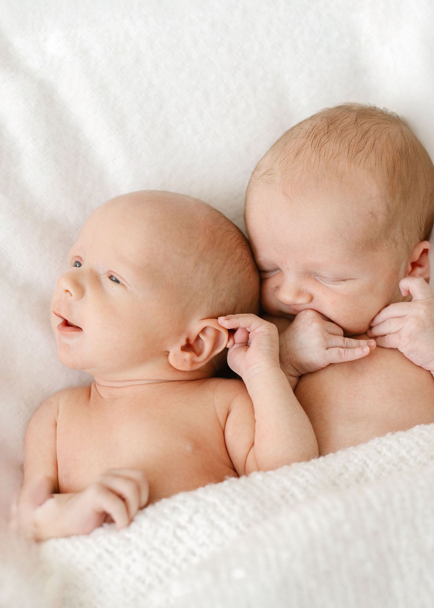 Two newborn twins lying together on a soft white blanket, one awake and one sleeping peacefully.