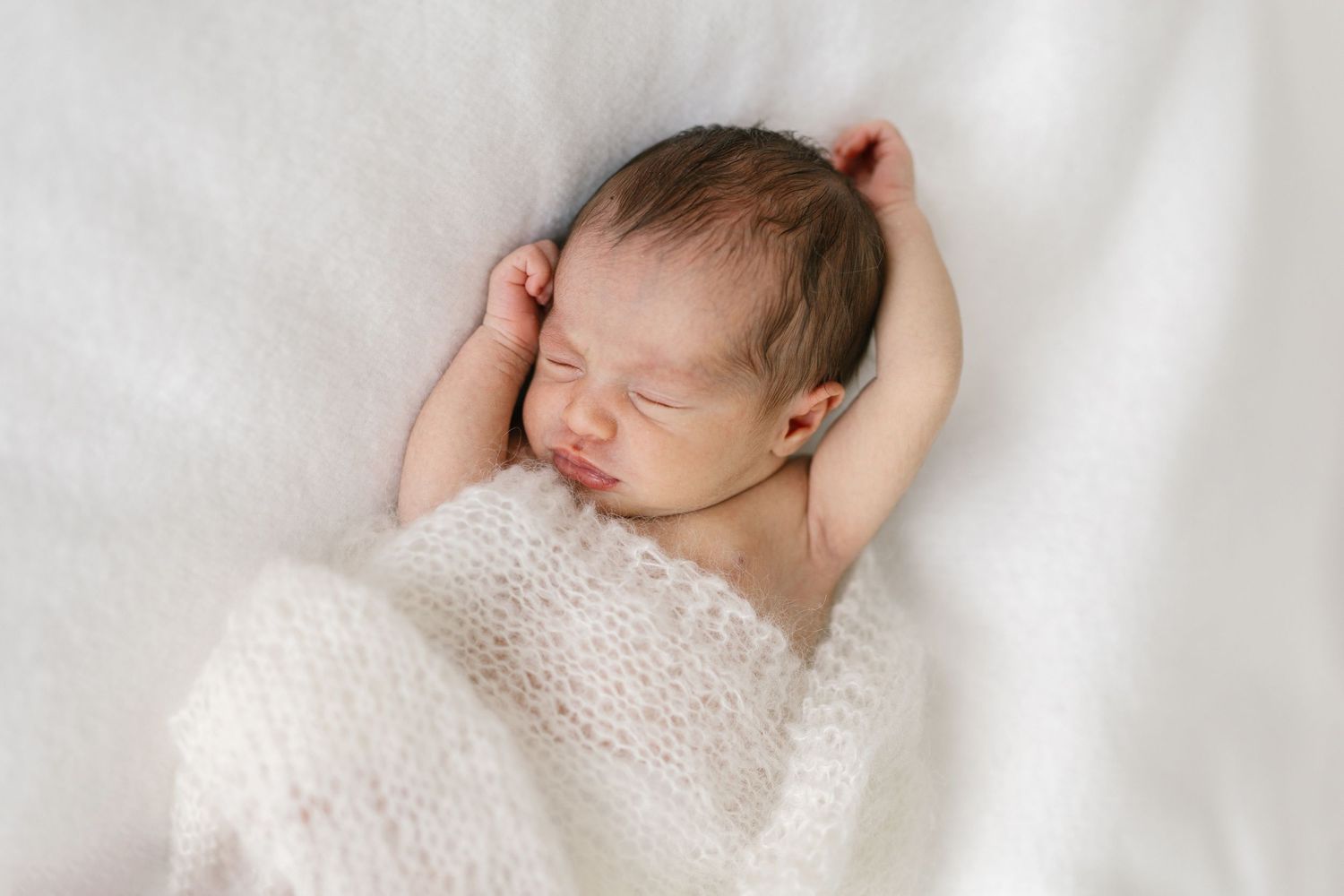 Peaceful newborn baby sleeping on white blanket with hands raised beside head, wrapped in soft knit wrap.