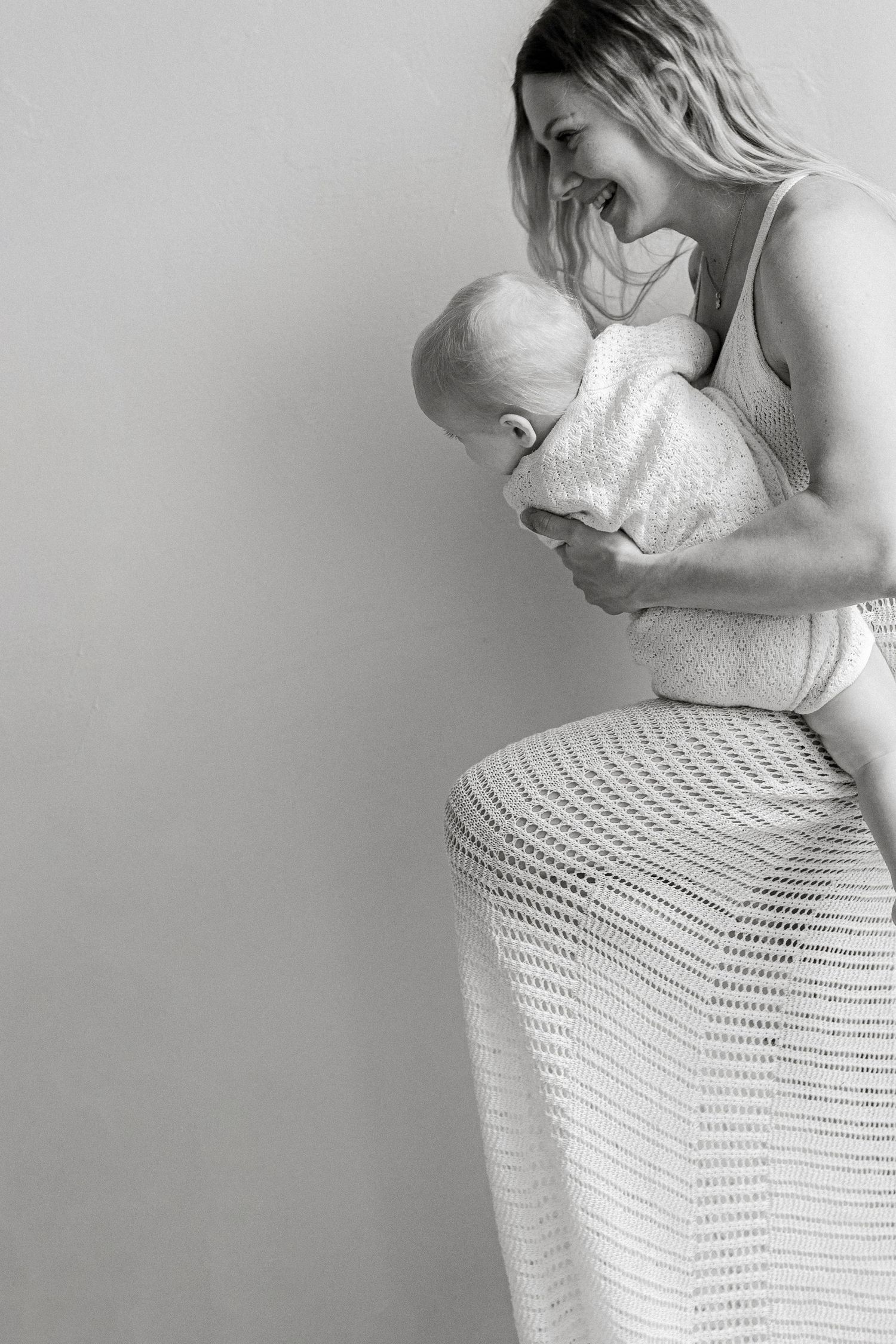 Black and white portrait of a smiling woman in a crochet skirt lifting one knee while tenderly cradling a baby in a knit outfit.