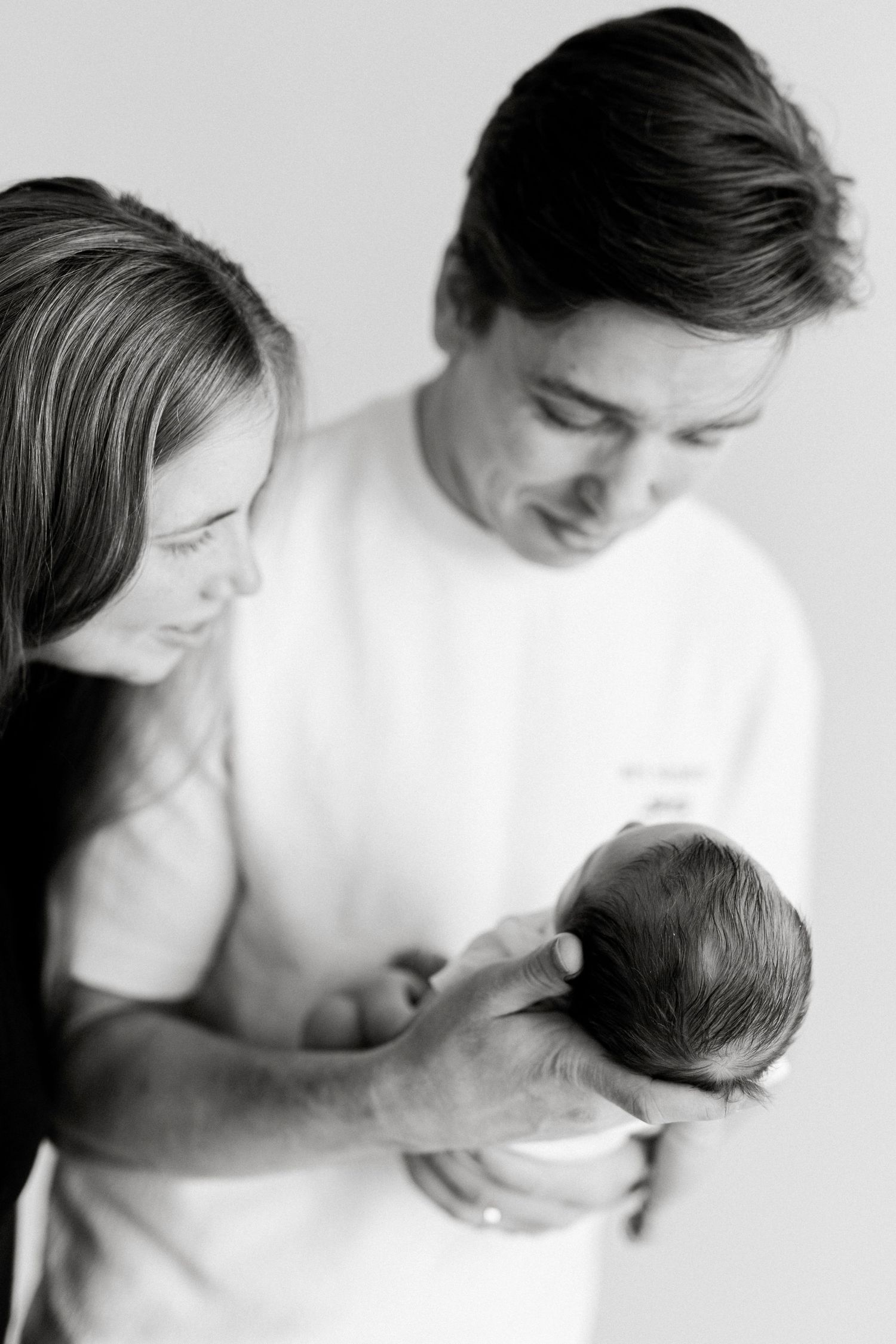 Black and white photo of parents lovingly gazing at their newborn baby held gently in fathers hands.