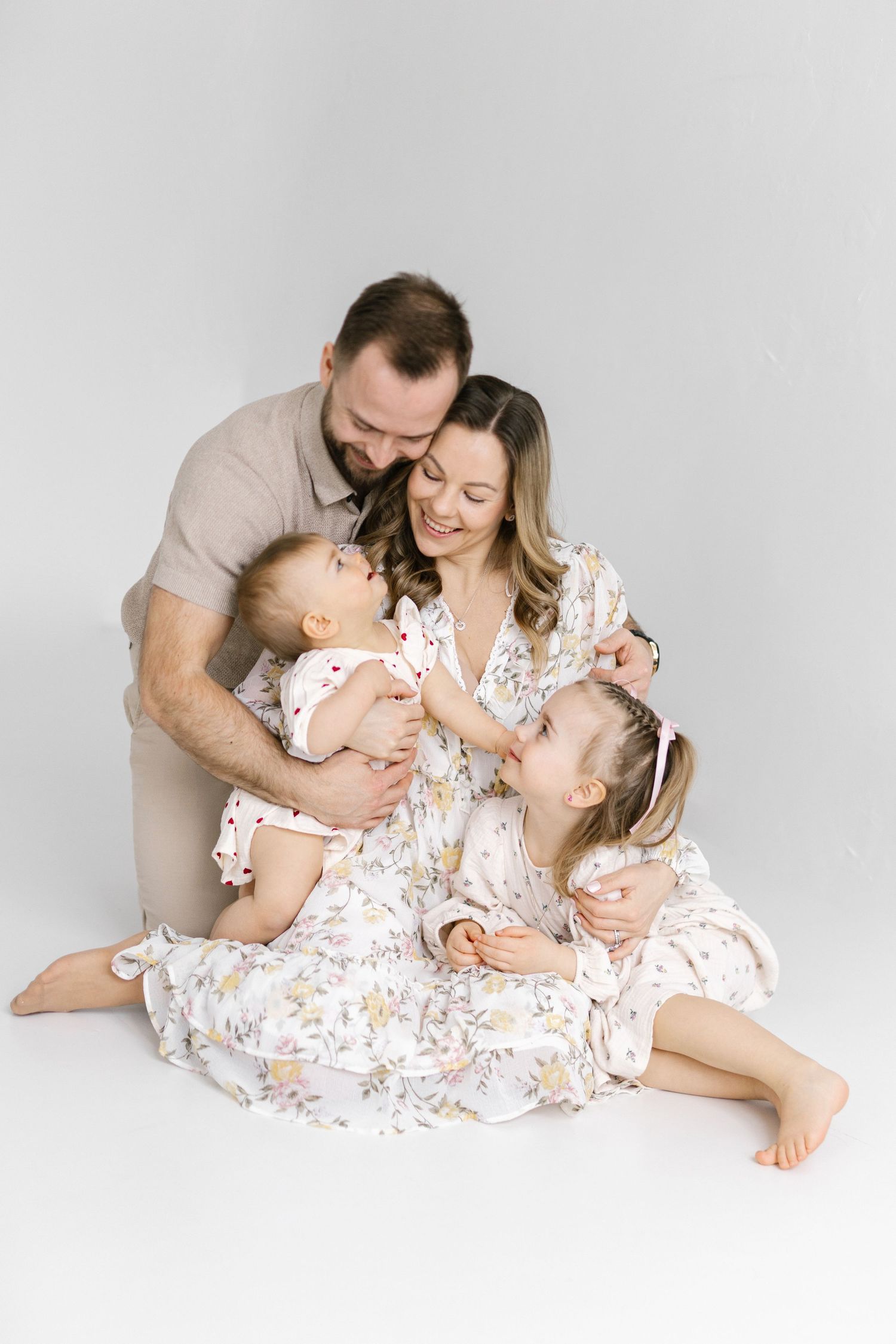 Happy family of four sitting together on white background, parents with two young daughters in floral dresses.