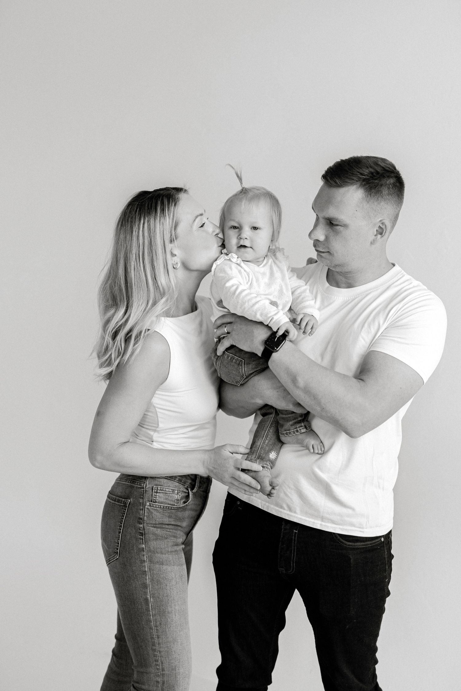 Black and white family portrait of parents in white tees and jeans, mother kissing baby held by father.
