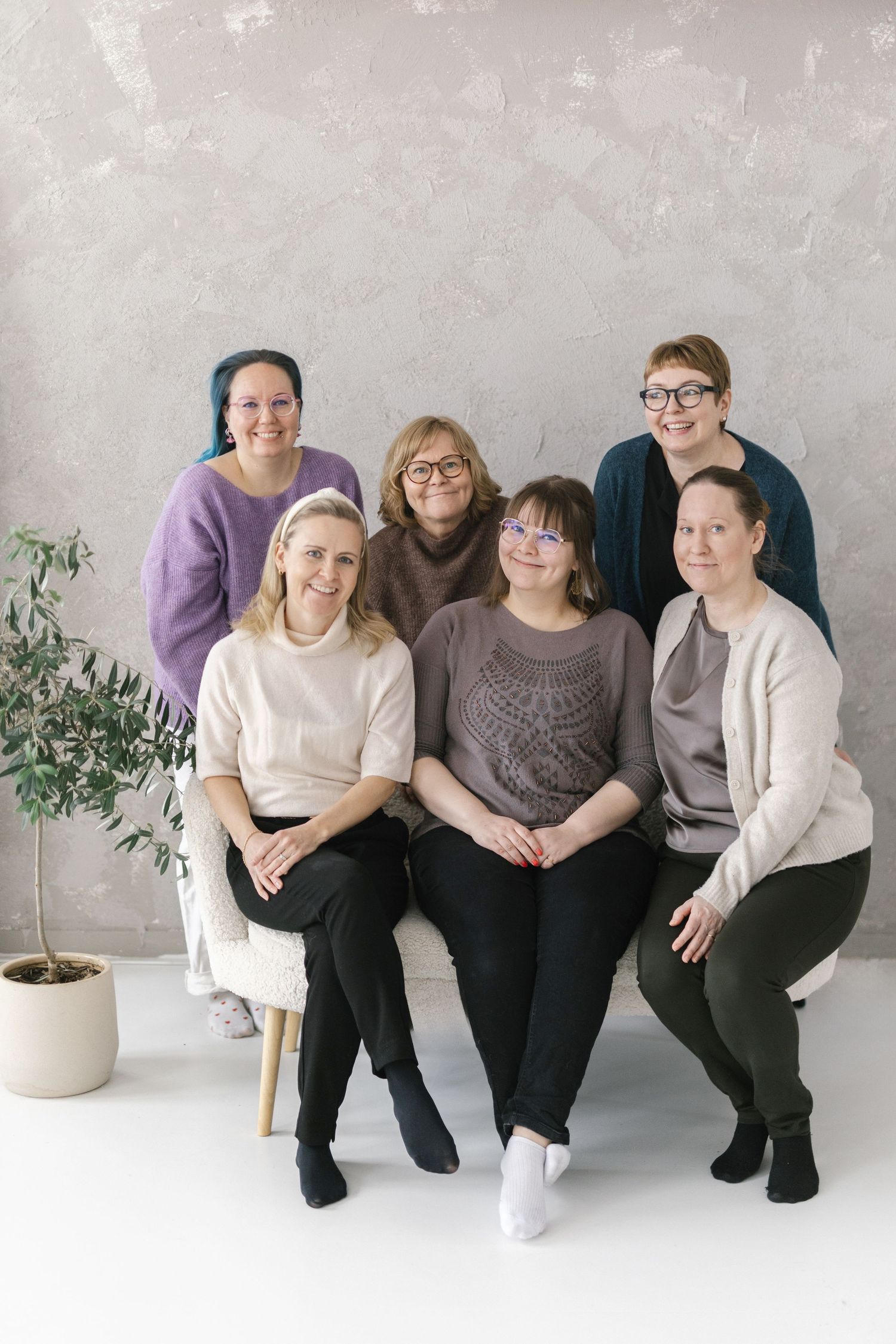 Six women posing together in a bright studio, three seated and three standing, wearing casual sweaters in neutral and purple tones.