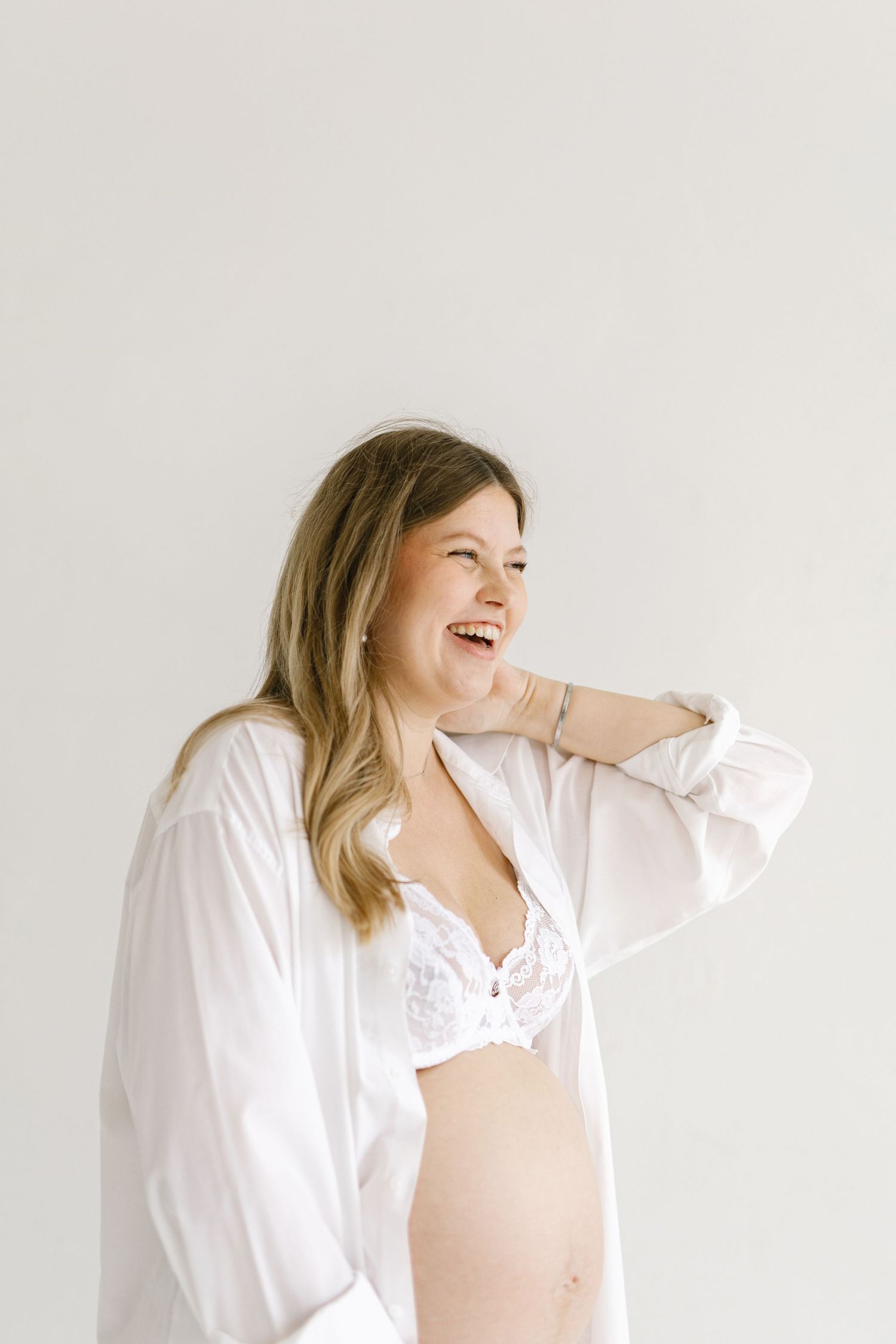Laughing pregnant woman in white shirt and lace bra, showcasing maternity fashion in a bright studio setting.