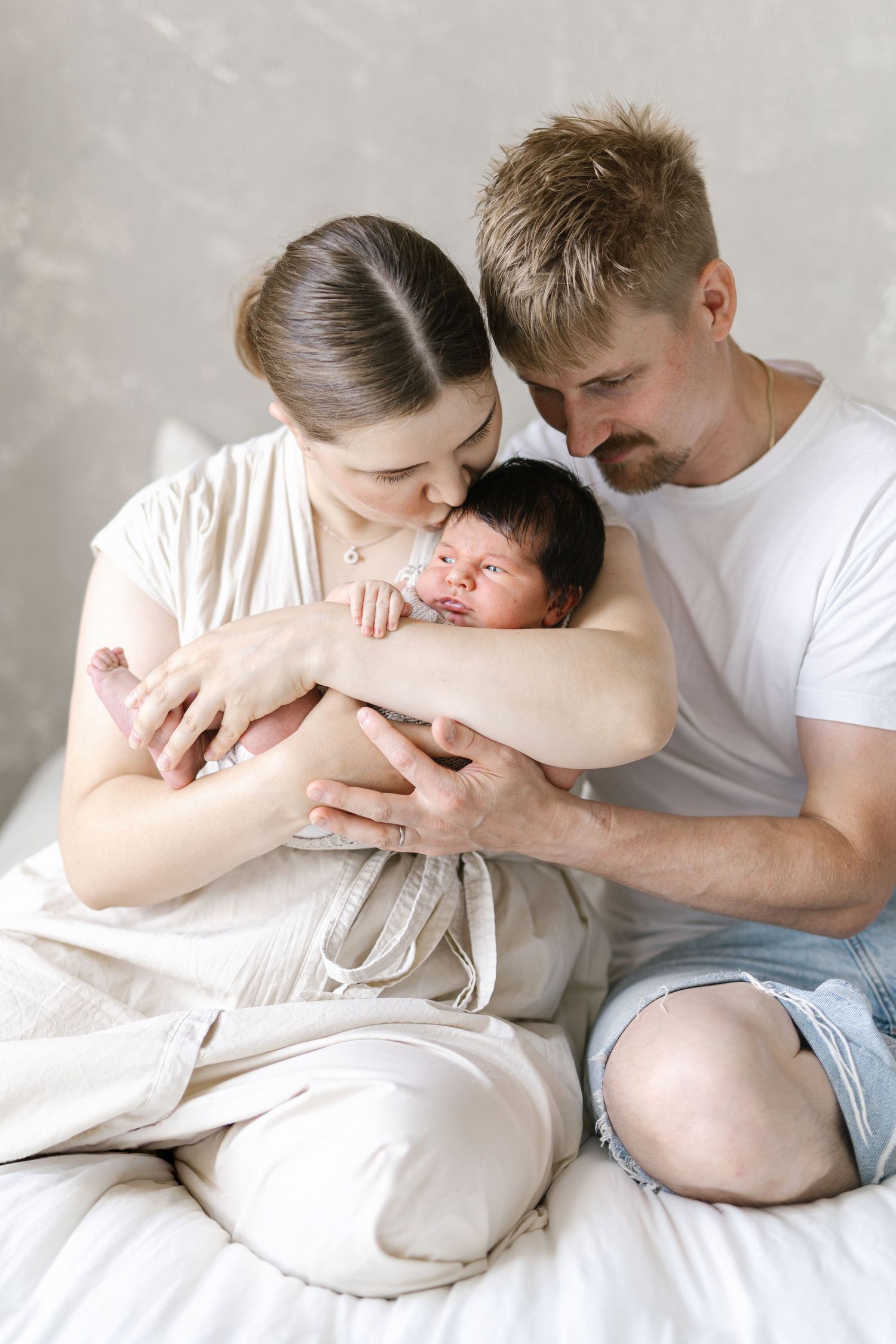 Young parents sitting on bed lovingly holding and kissing their newborn baby wrapped in white cloth.