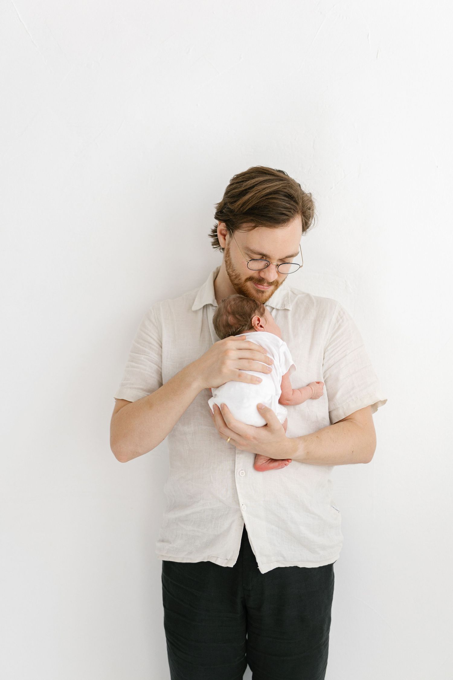 A young bearded father with glasses tenderly holds his newborn baby wrapped in white against a clean white background.