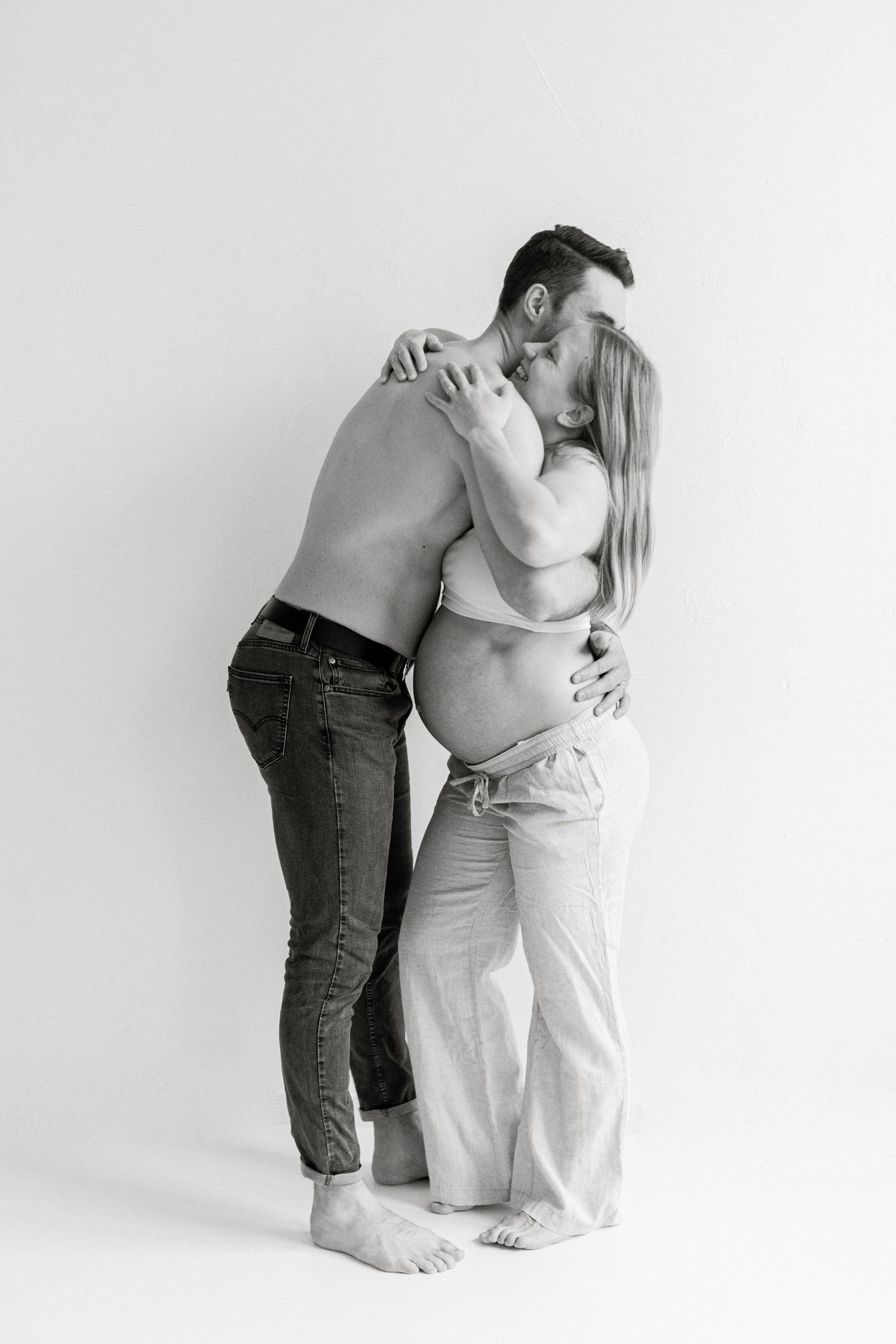 Black and white photo of a shirtless man kissing his pregnant partner while she cradles her baby bump.