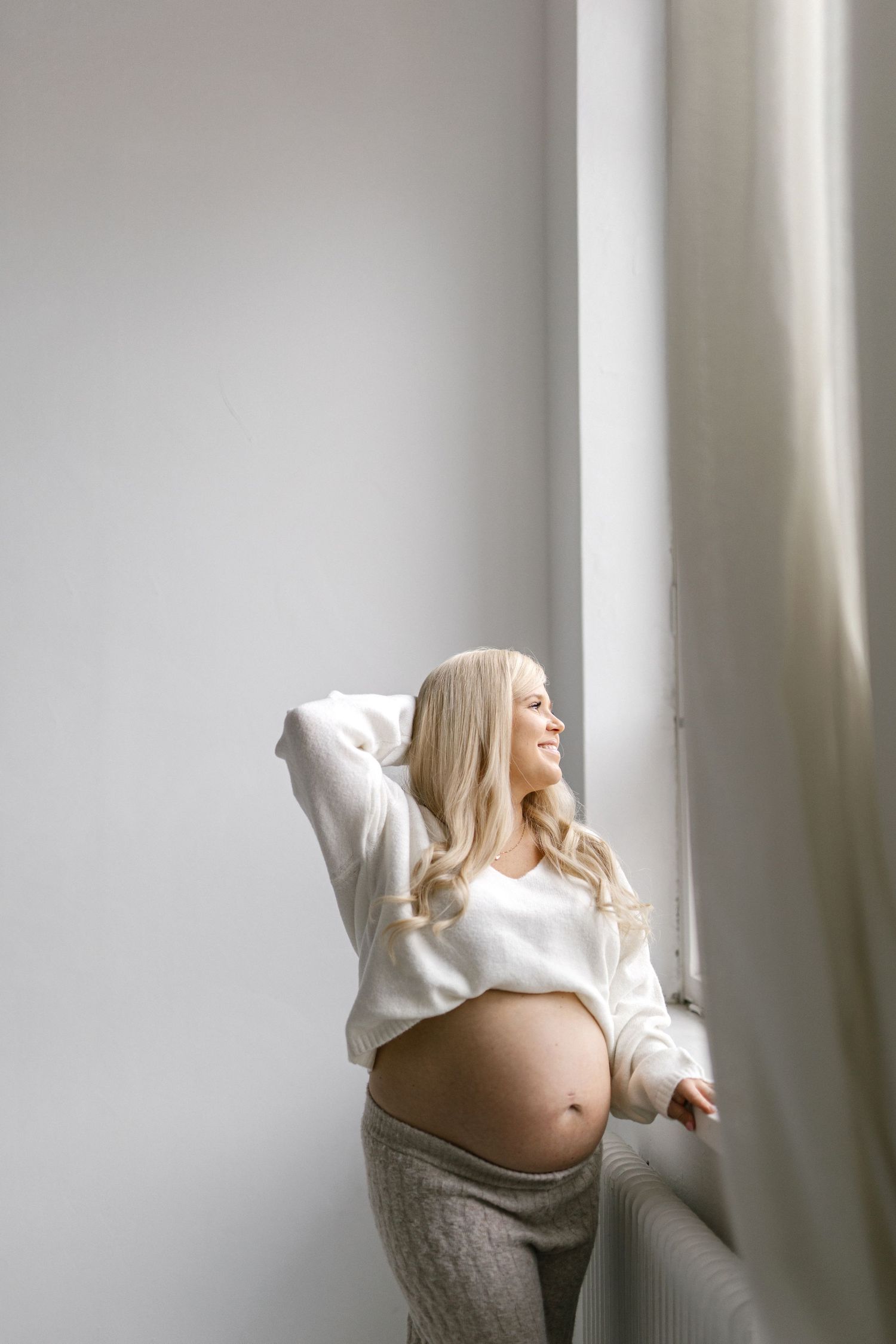 Smiling pregnant blonde woman in white crop top stands by window showing baby bump in bright minimalist room.