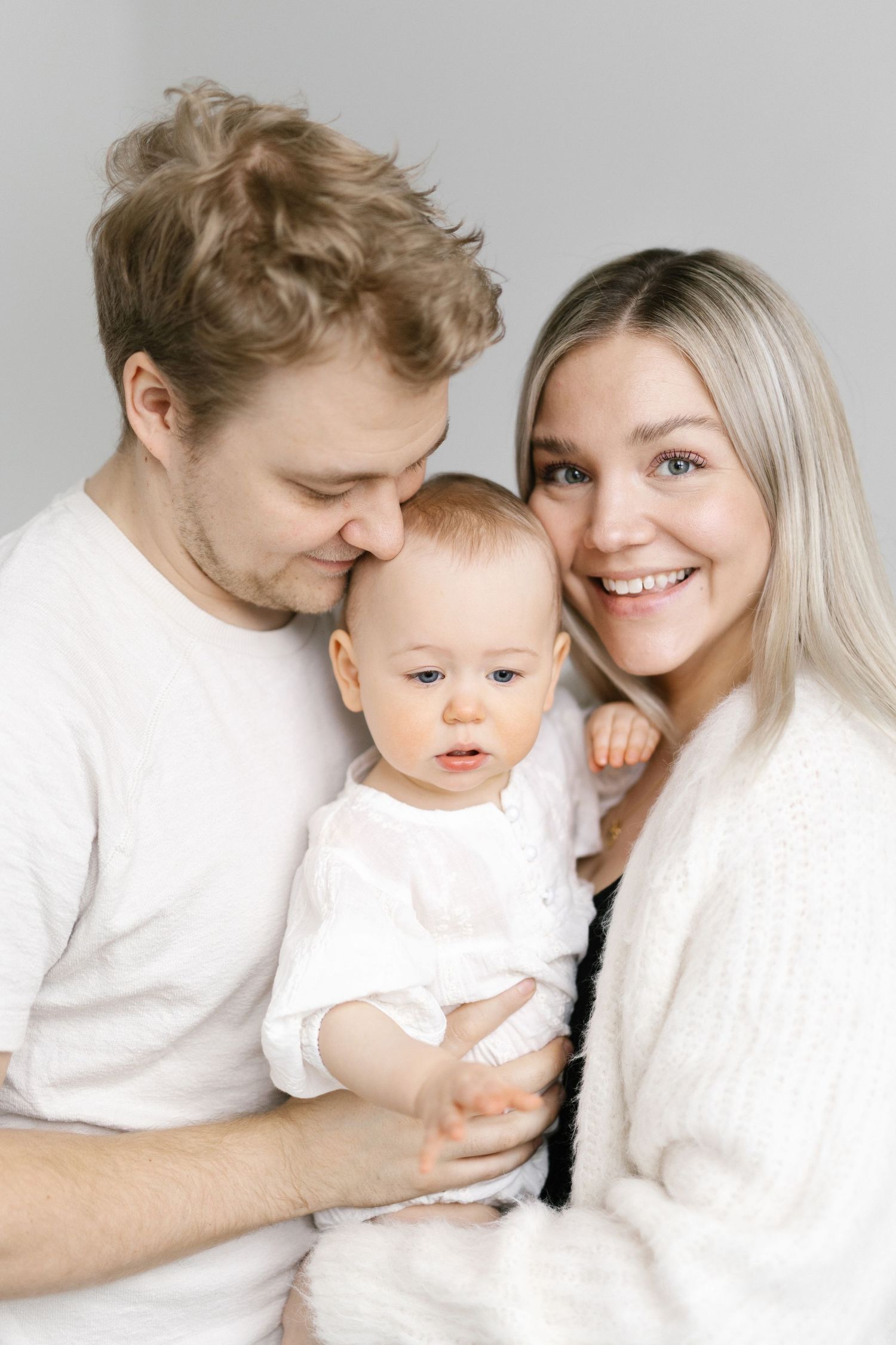 Happy young family with parents holding their smiling baby dressed in white against a light background.