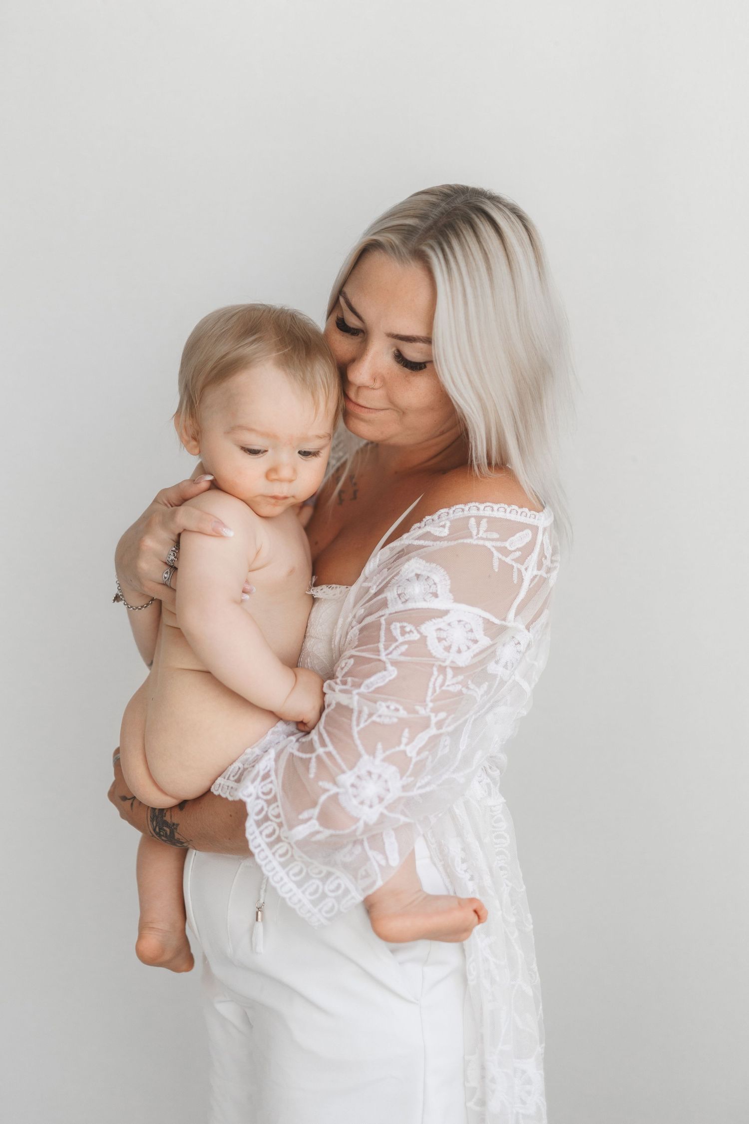 A blonde woman in a white lace dress tenderly holds a baby against a light background.