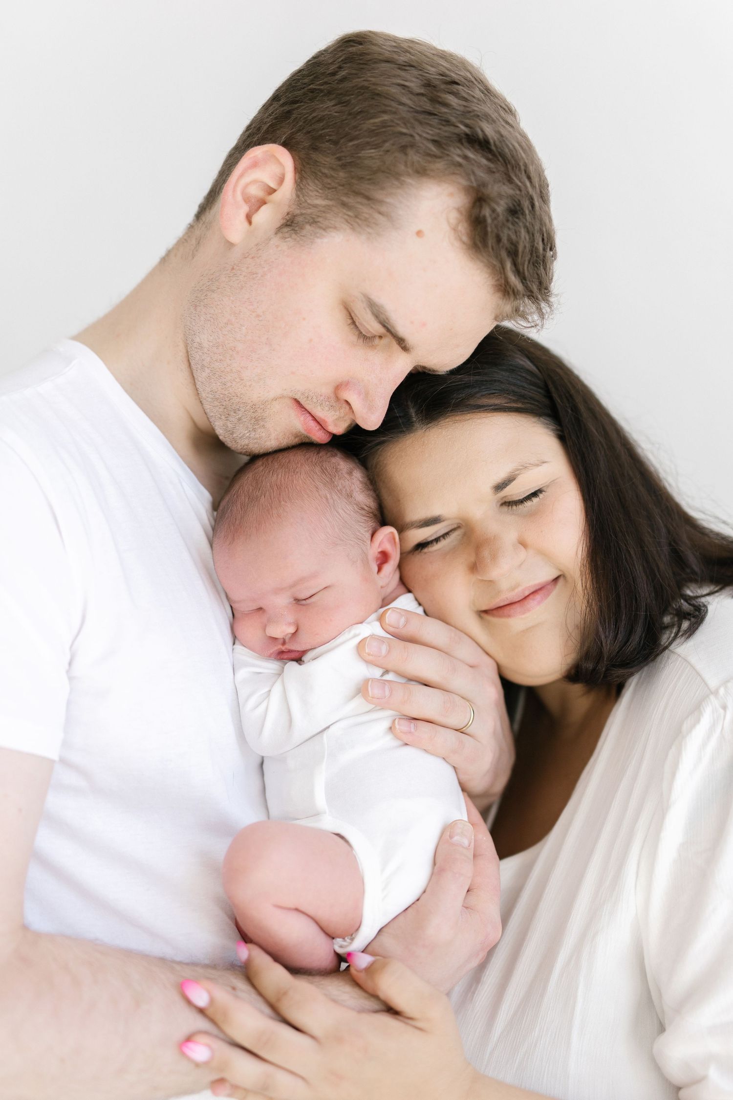 New parents tenderly holding their sleeping newborn baby, all dressed in white, in a heartwarming family portrait.