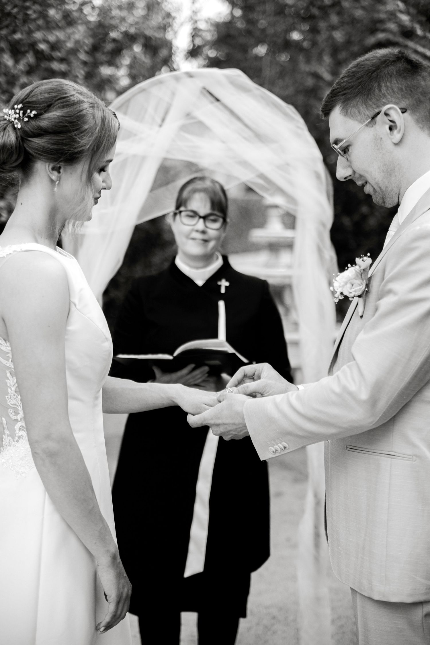 Black and white wedding ceremony photo of bride and groom exchanging rings before a female officiant holding a bible.