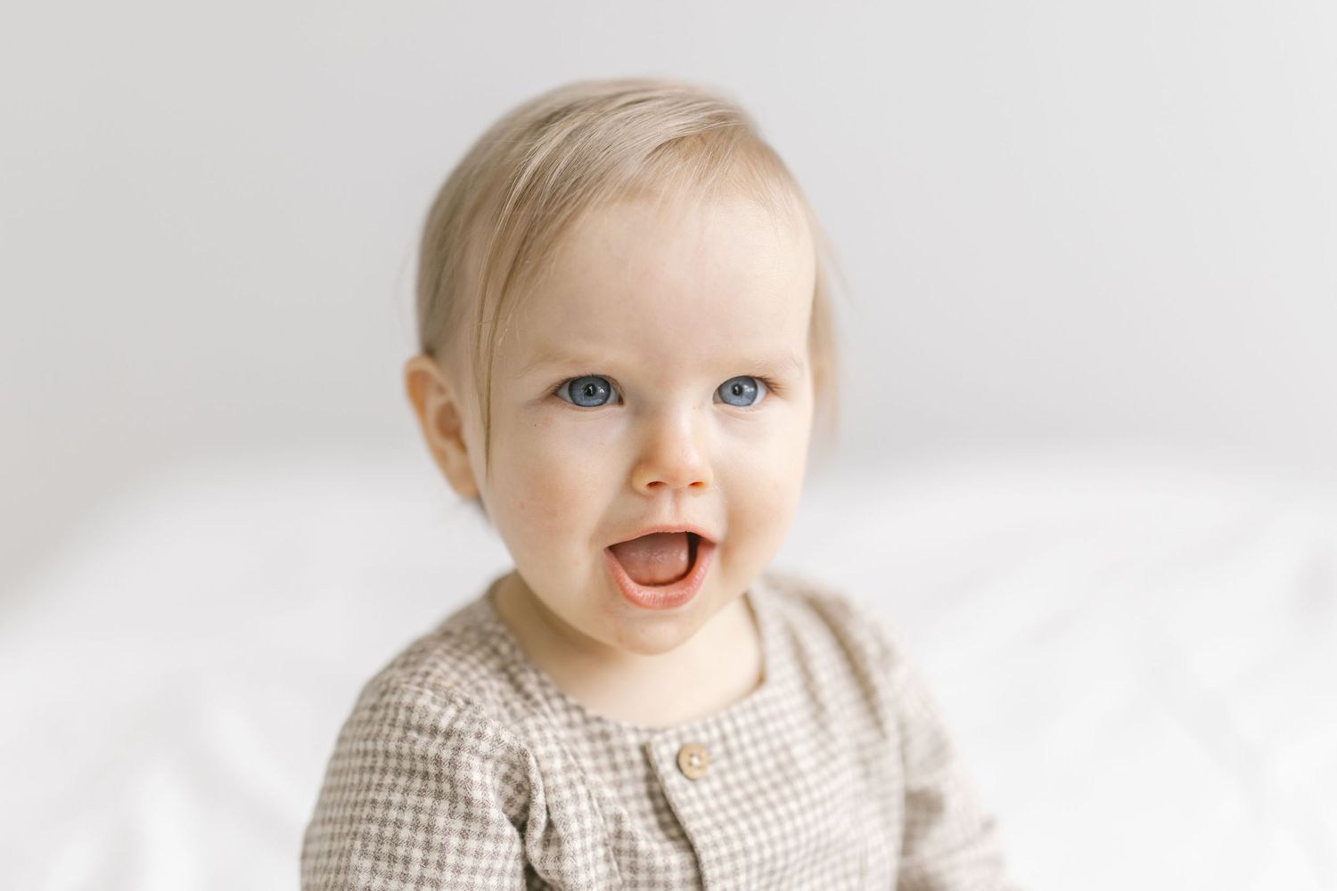 Baby wearing gingham dress smiles sweetly against white background in studio portrait series.