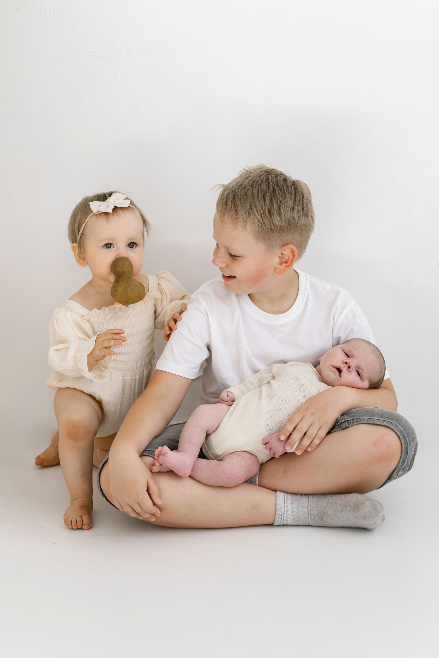 Three siblings sitting together with toddler holding pacifier and newborn baby.