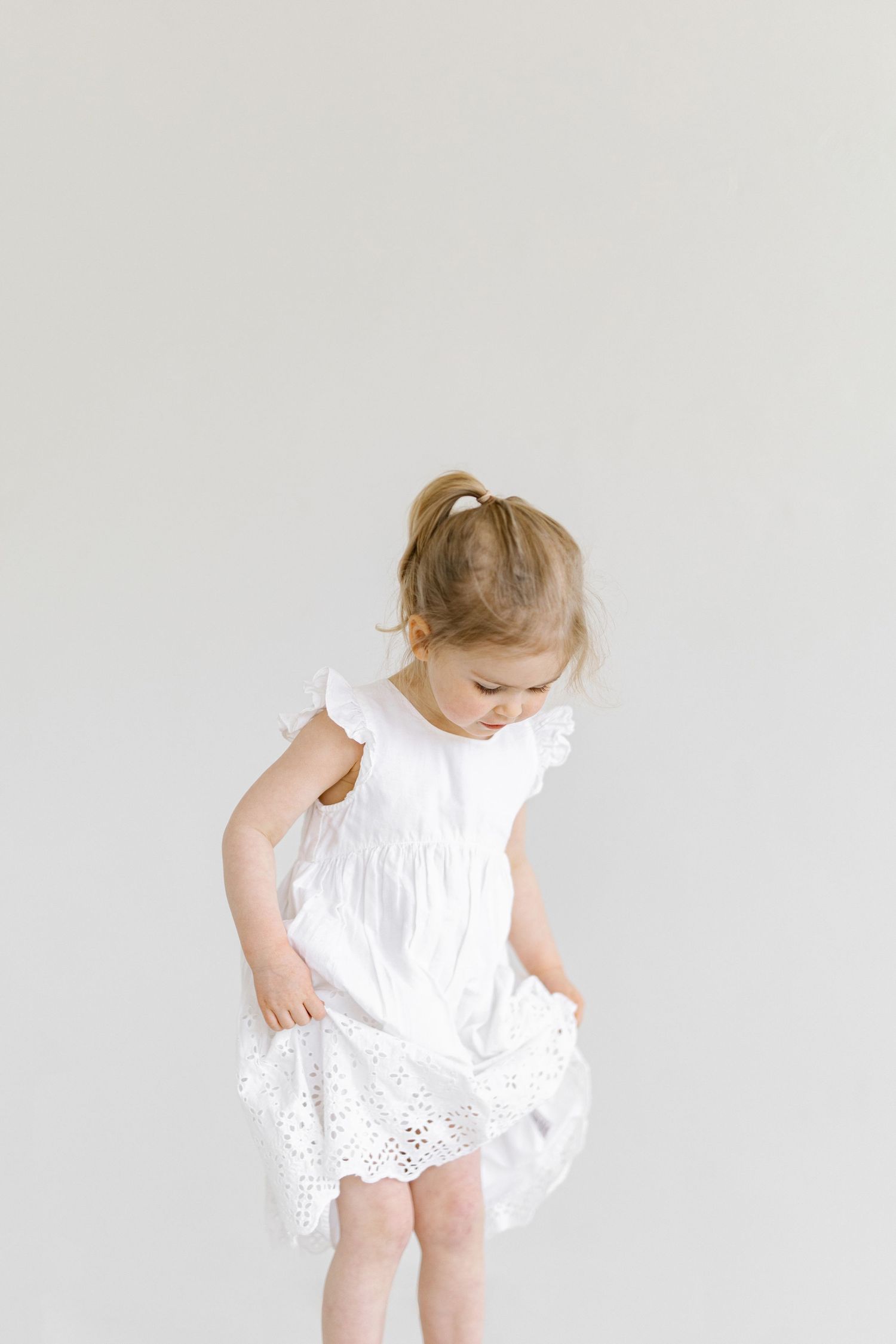 Young girl in white eyelet dress twirling, looking down, against light gray background.