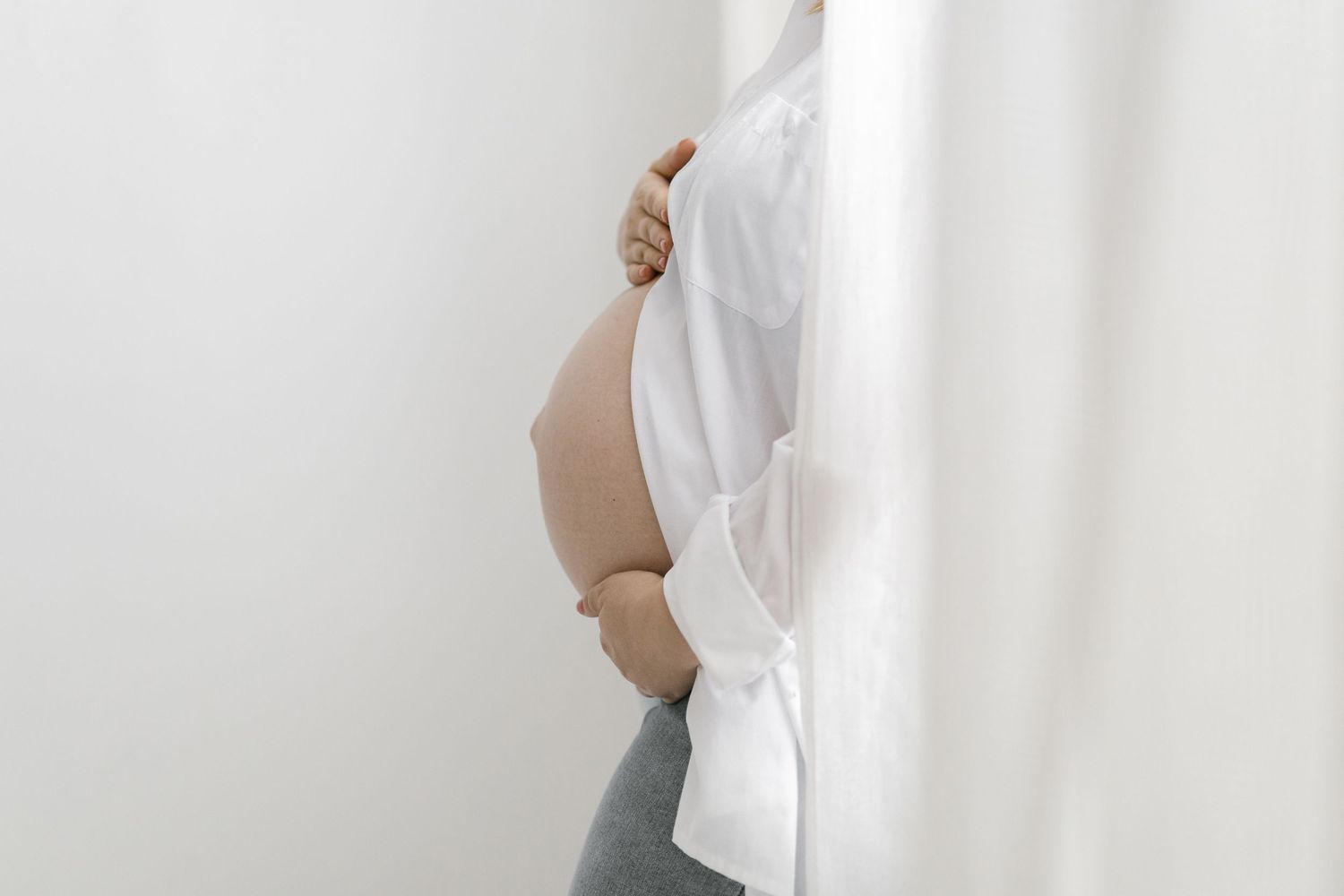 Pregnant woman in white shirt cradling baby bump against white wall, side profile maternity photo.