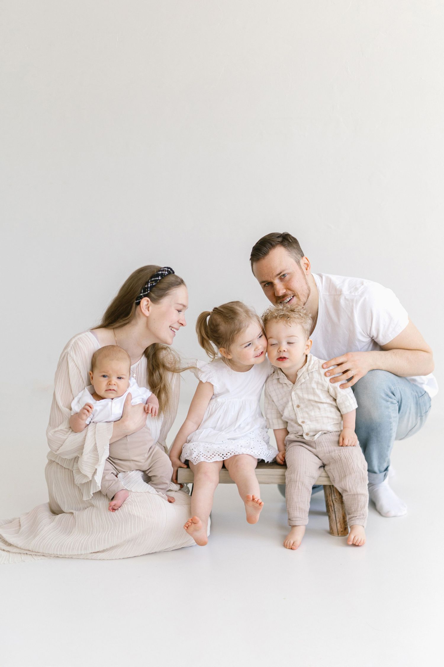A happy young family with three small children poses together in a bright white studio for a professional portrait.