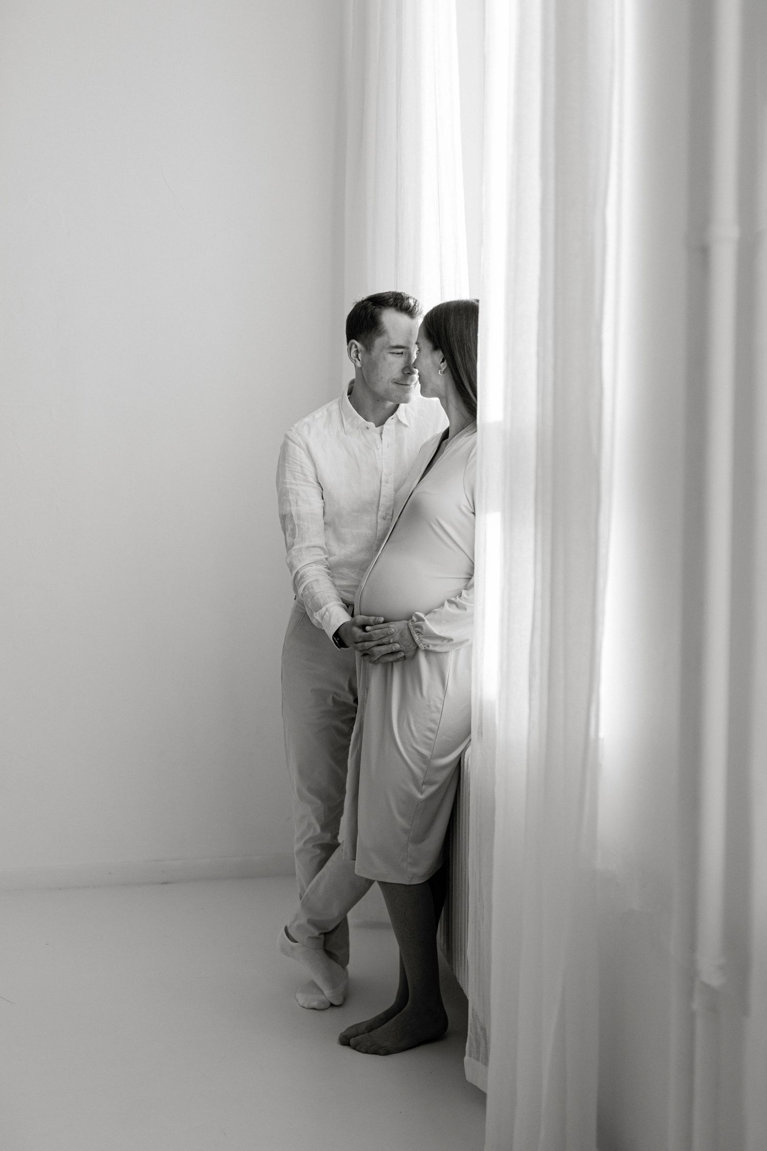 Black and white photo of a couple sharing a tender moment by sheer curtains, the woman visibly pregnant.