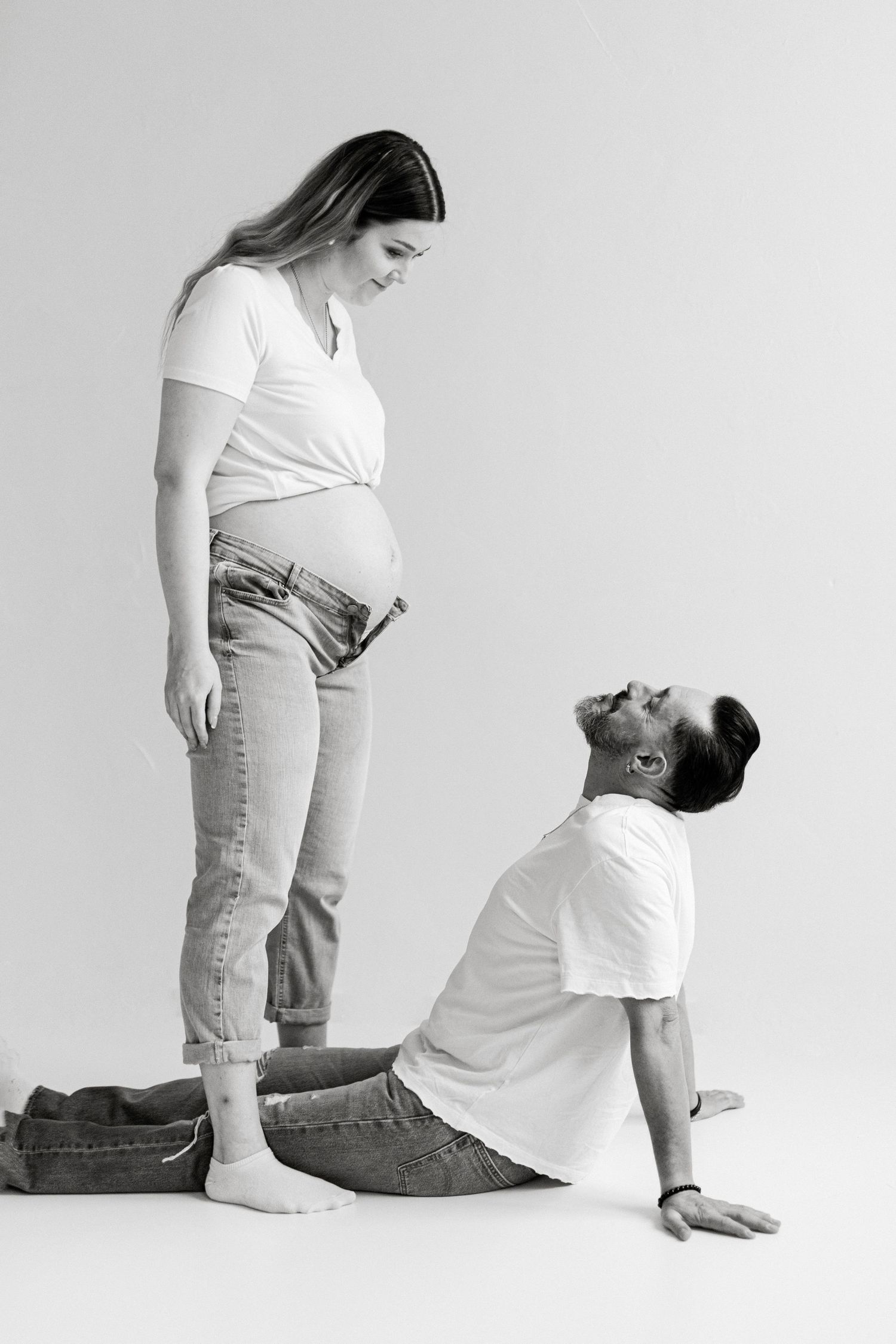 Black and white photo of a pregnant woman standing while her partner sits on the floor gazing up at her baby bump.