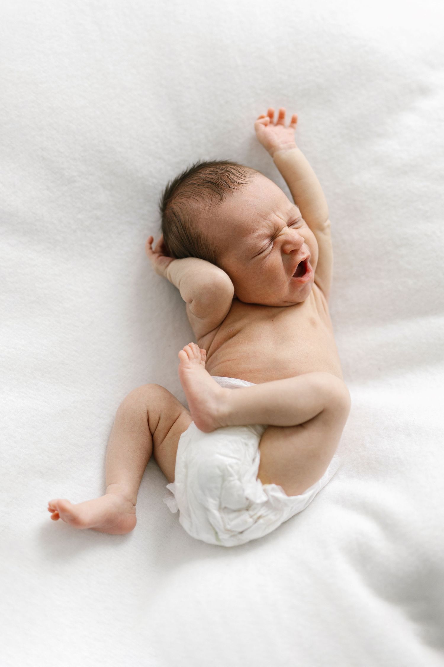 Newborn baby in a white diaper lying on a white blanket, yawning and stretching arms upward.