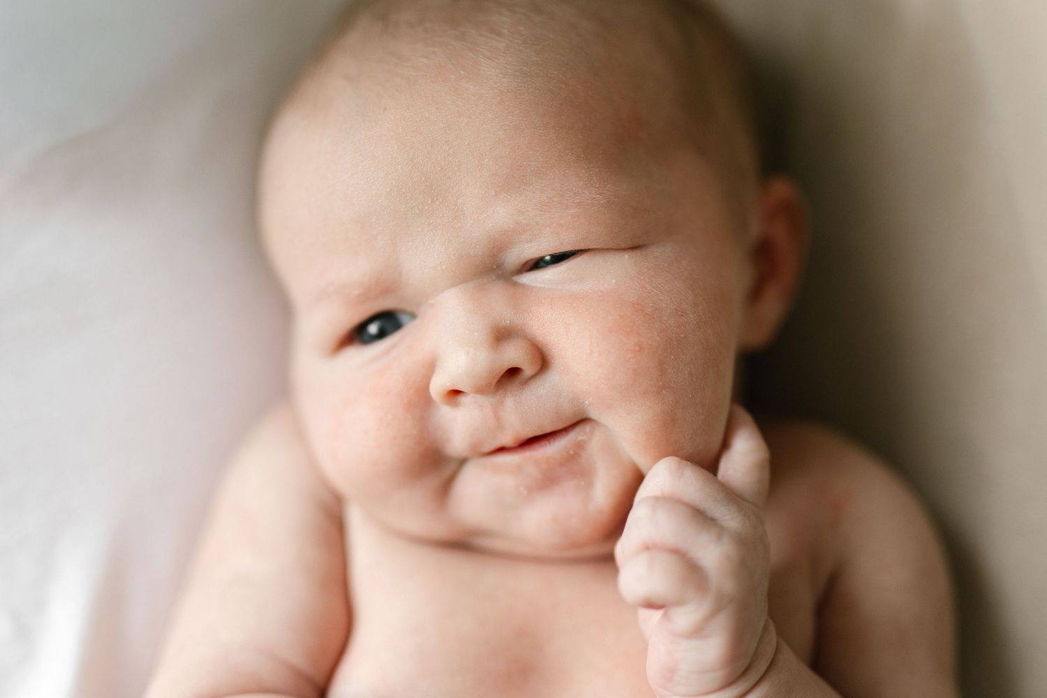 Adorable newborn baby with chubby cheeks resting finger on chin, making a cute squinting expression.