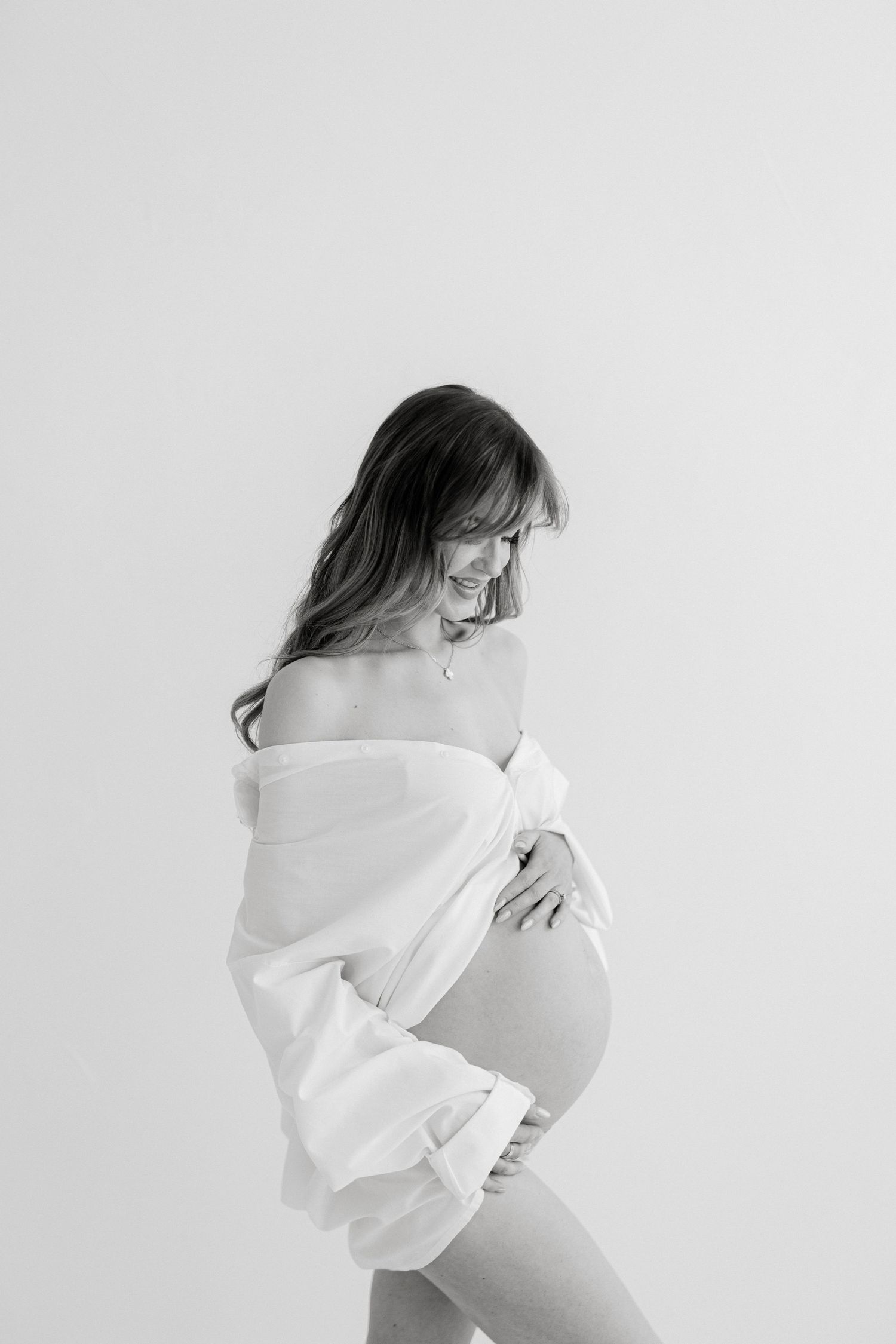 Black and white maternity photo of a smiling pregnant woman wrapped in a white fabric, cradling her bare baby bump.