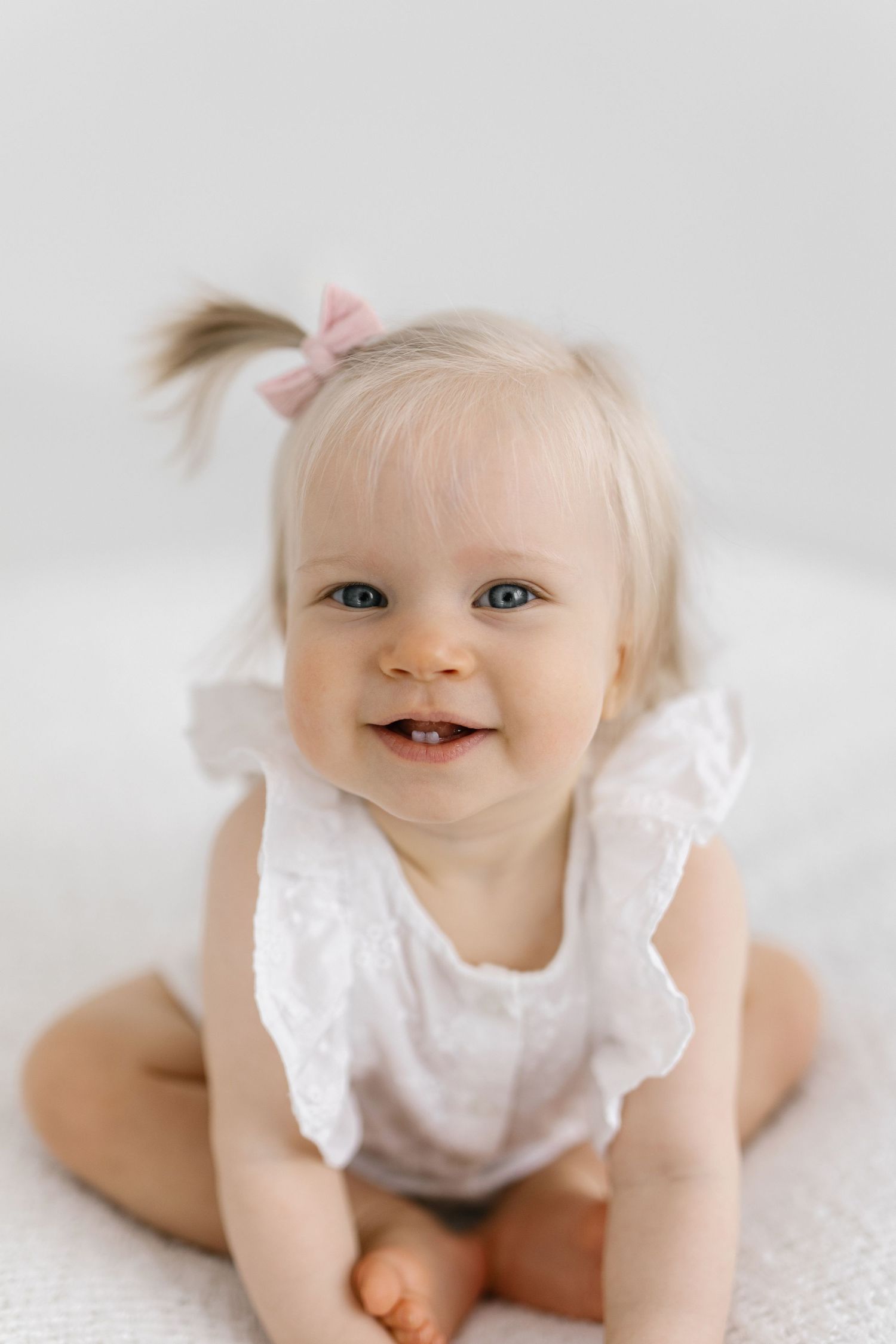 Baby in white lace dress lying on white surface with joyful expression.