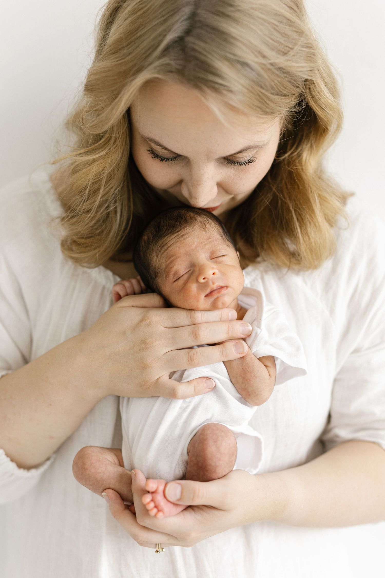 A blonde mother in white gently kisses her sleeping newborn baby dressed in a white onesie.