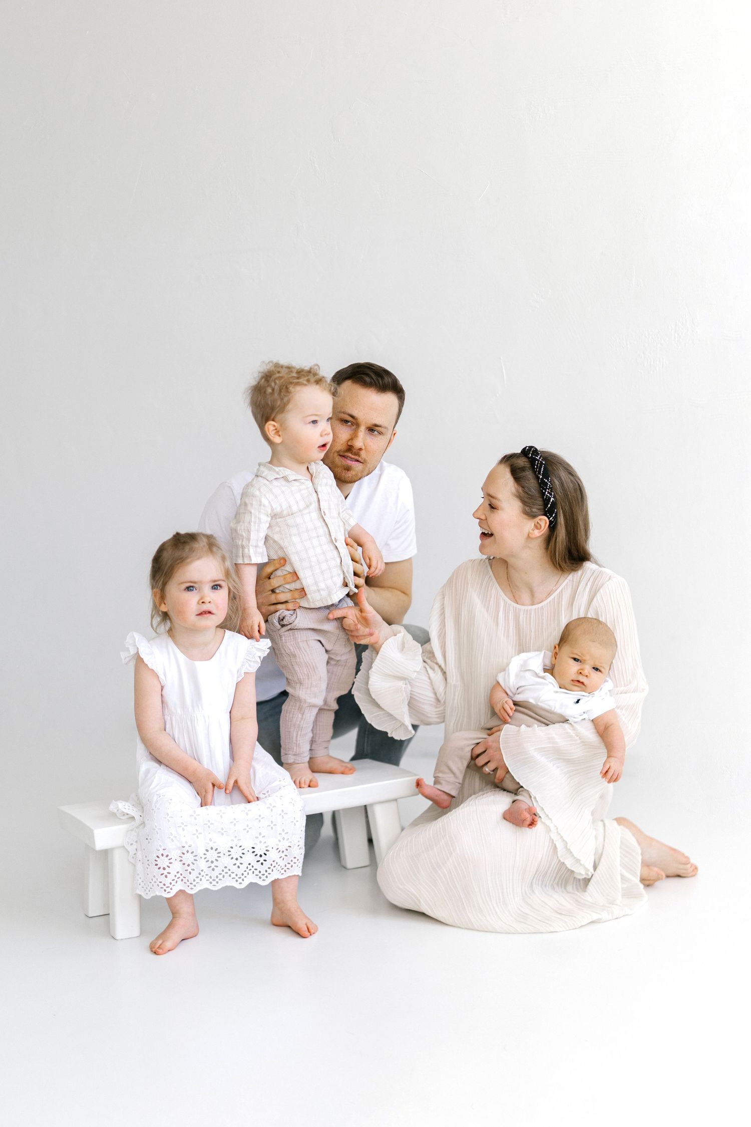 A young family with three children poses together in white outfits against a white background in a studio portrait.