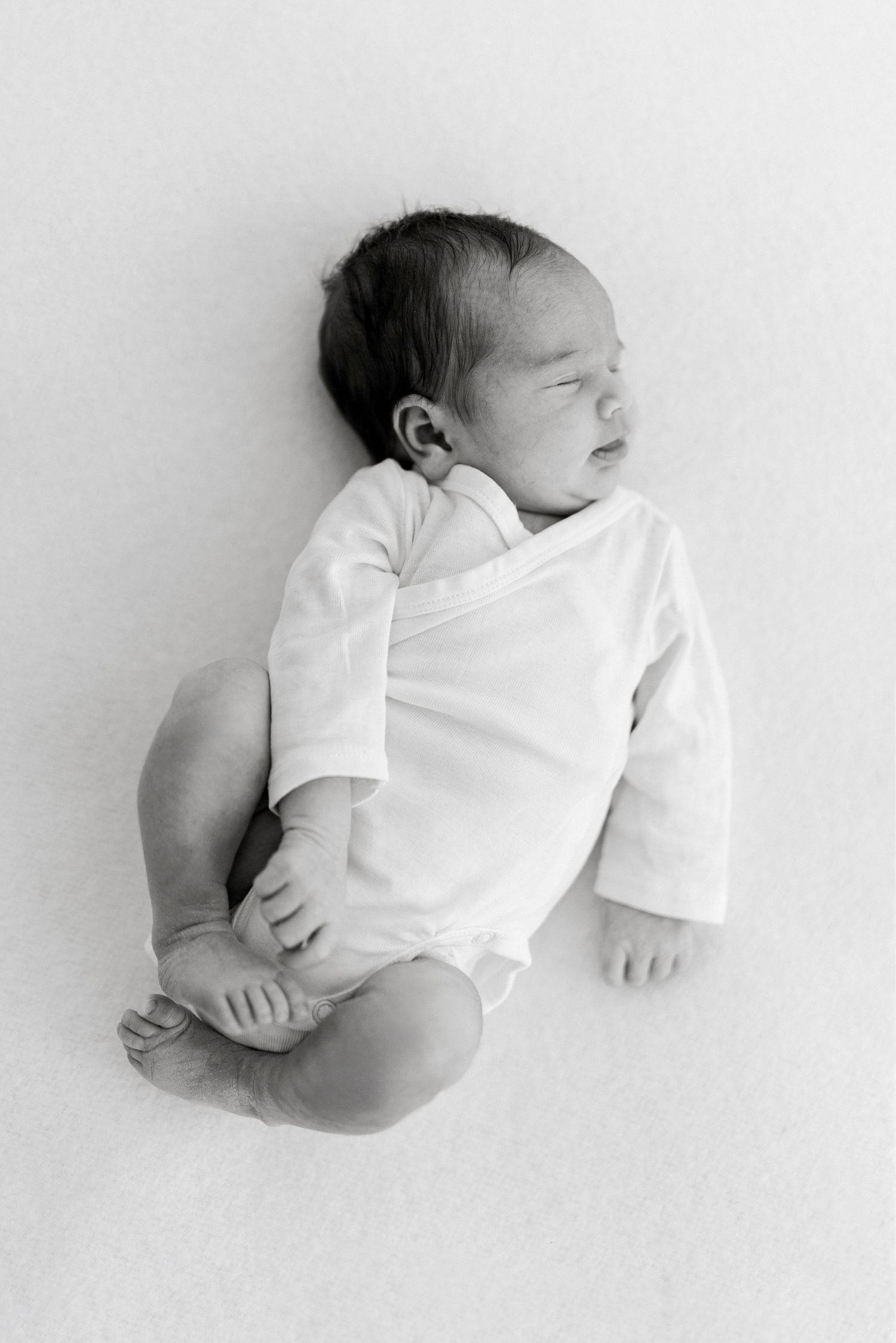A sleeping newborn baby wearing a white onesie lies peacefully against a light background in black and white.
