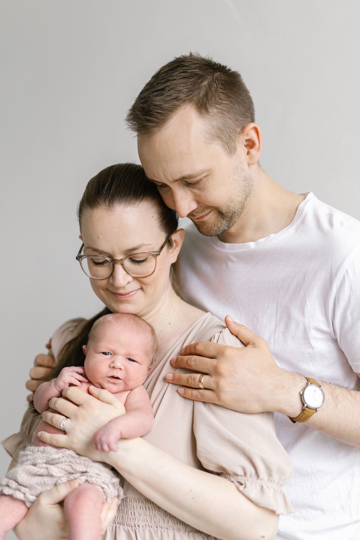 Young parents lovingly holding their newborn baby against a soft gray background in a tender family portrait.