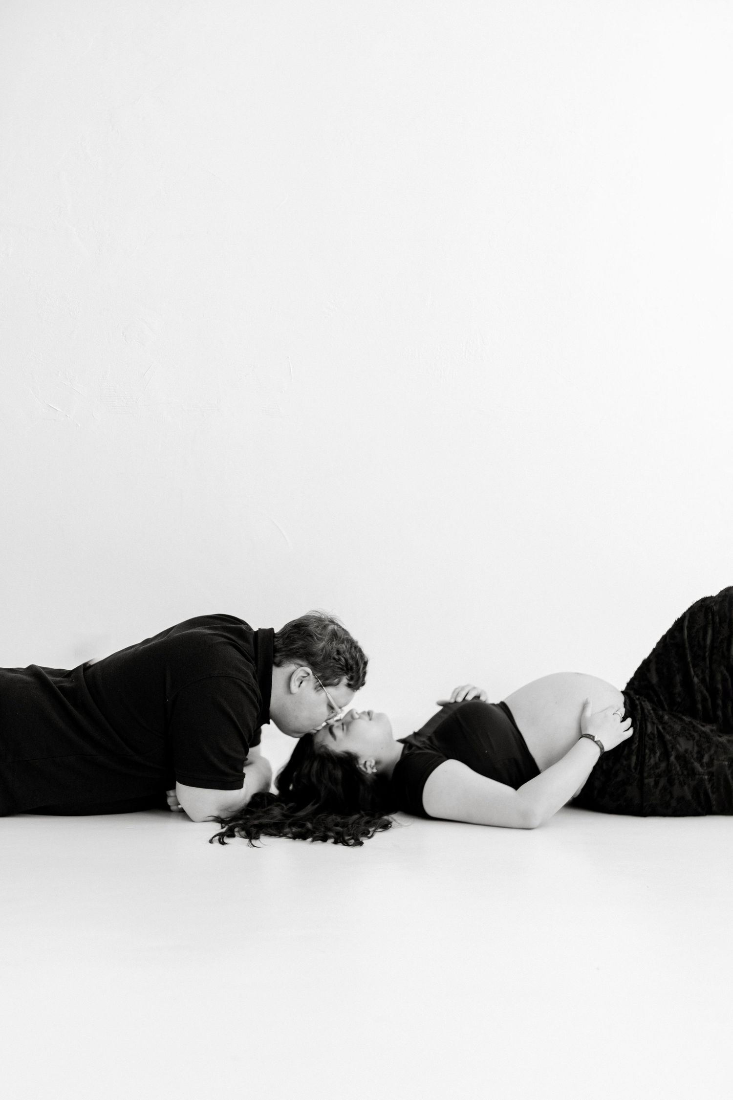 Black and white photo of a couple sharing a kiss while lying on a white studio floor, woman in a black dress.