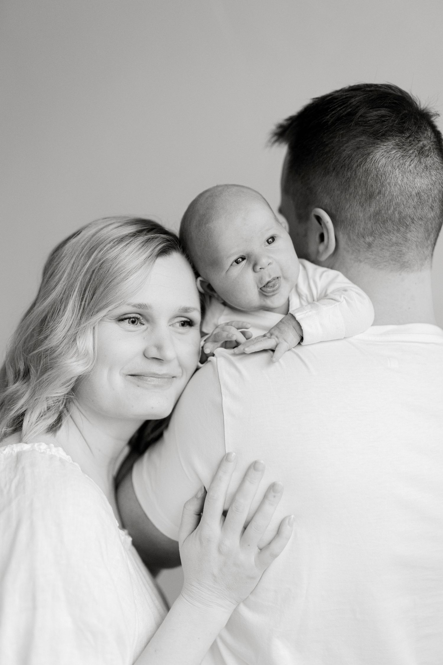 Parents in white sharing tender moments with baby against light background.