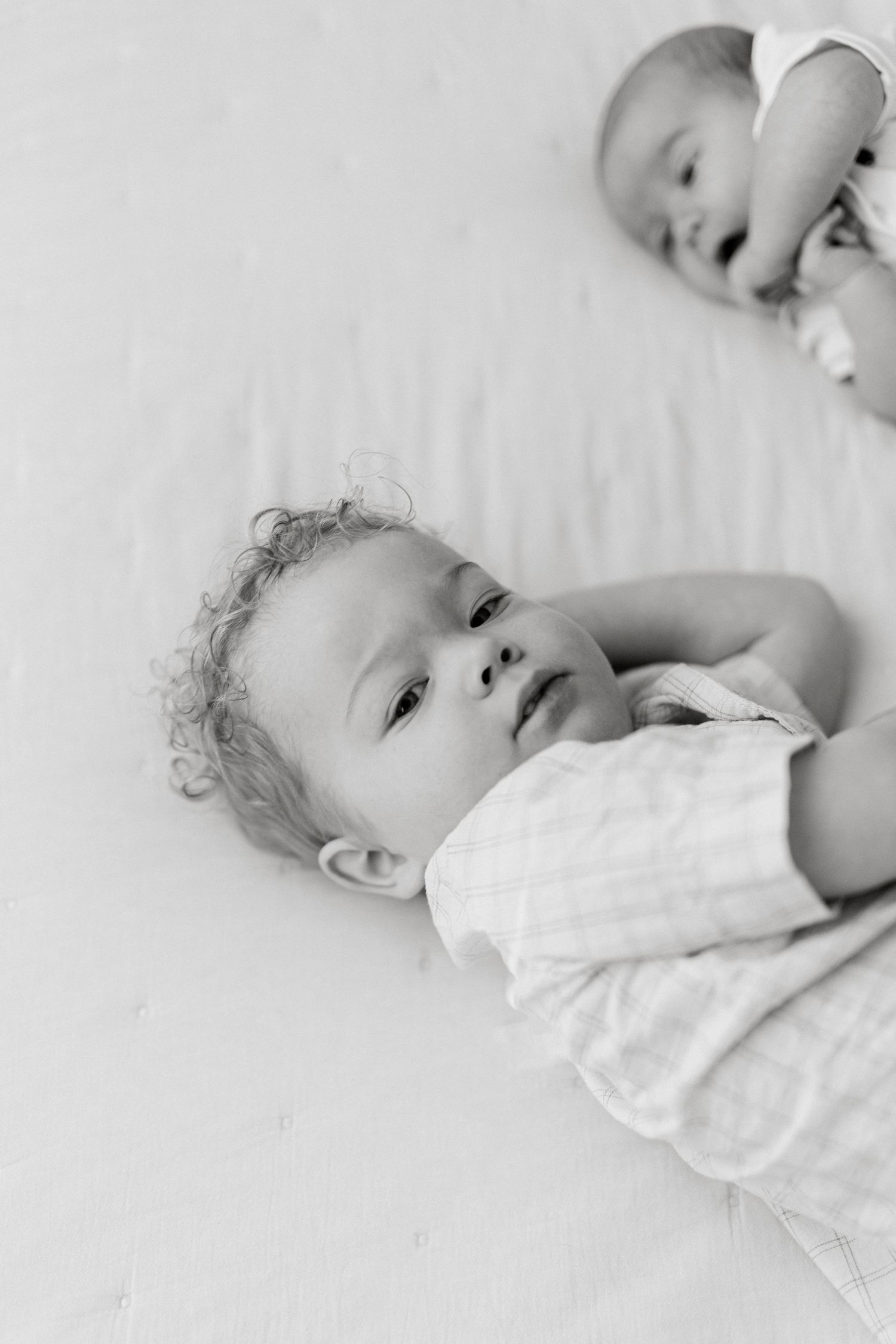 Black and white photo of two newborn babies lying on a soft white blanket, one with curly hair looking up.