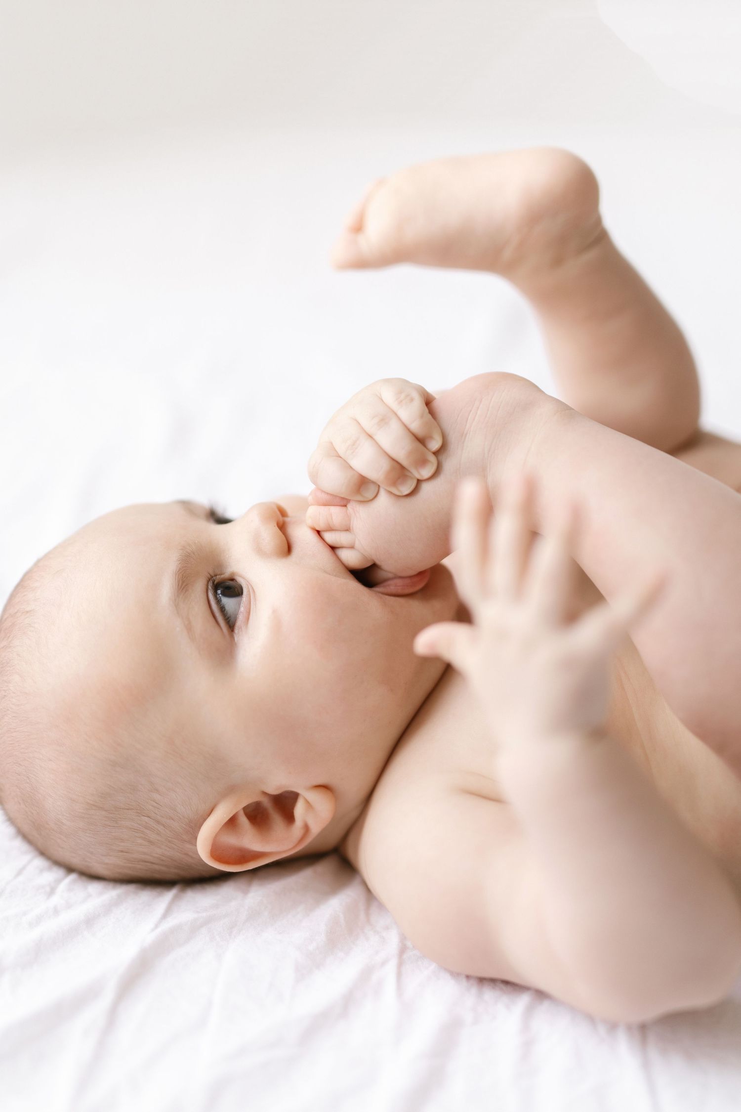 Cute naked baby lying on white blanket, holding foot with hands, looking up with curious expression.
