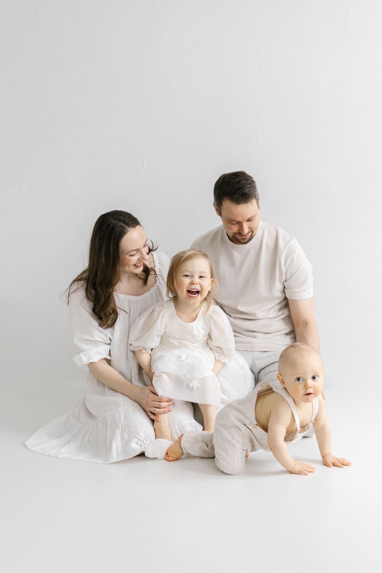 Happy family in white outfits sitting on floor, parents smiling at laughing toddler while baby crawls nearby.