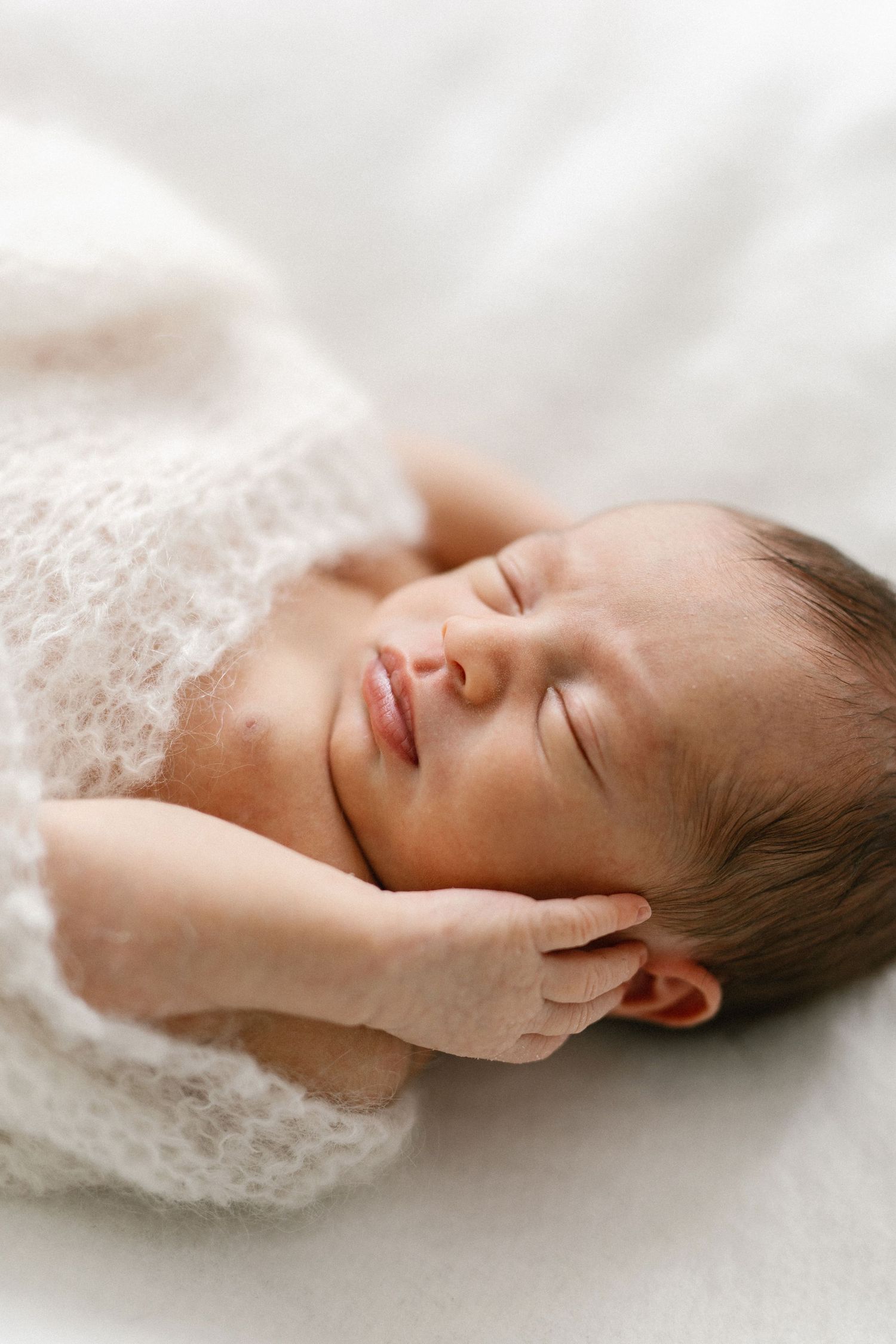 Peaceful newborn baby sleeping wrapped in white knit blanket with tiny hand resting near face.