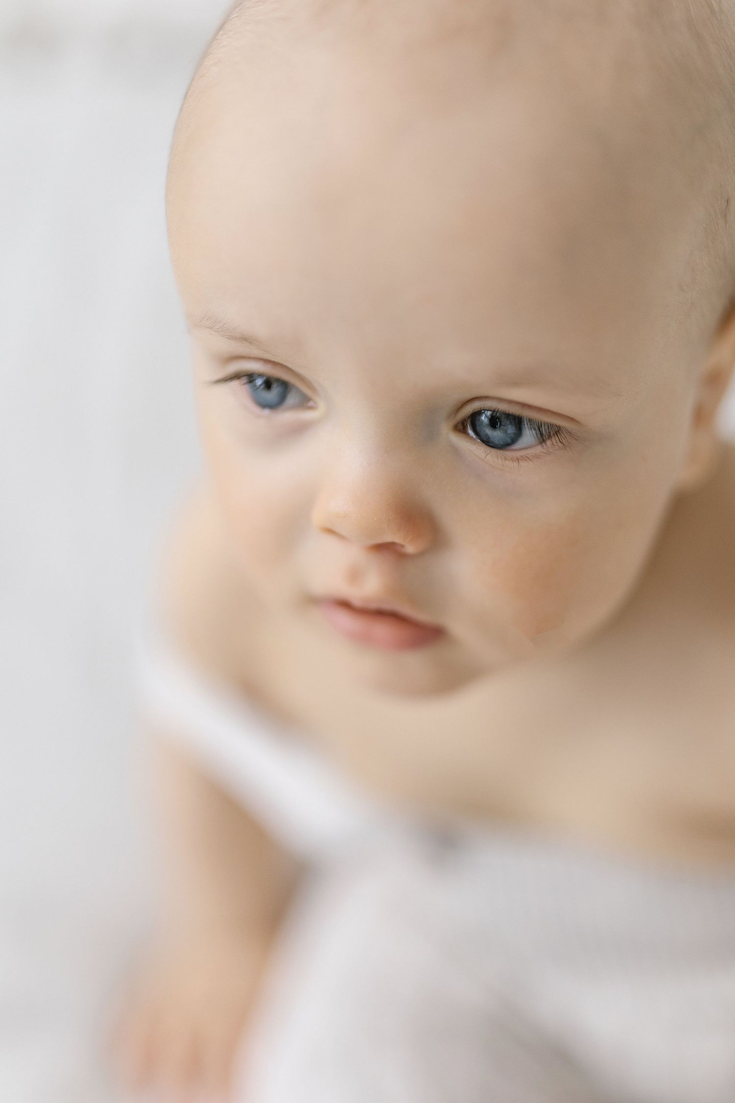 Close-up portrait of a cute baby with striking blue eyes and fair skin against a soft white background.
