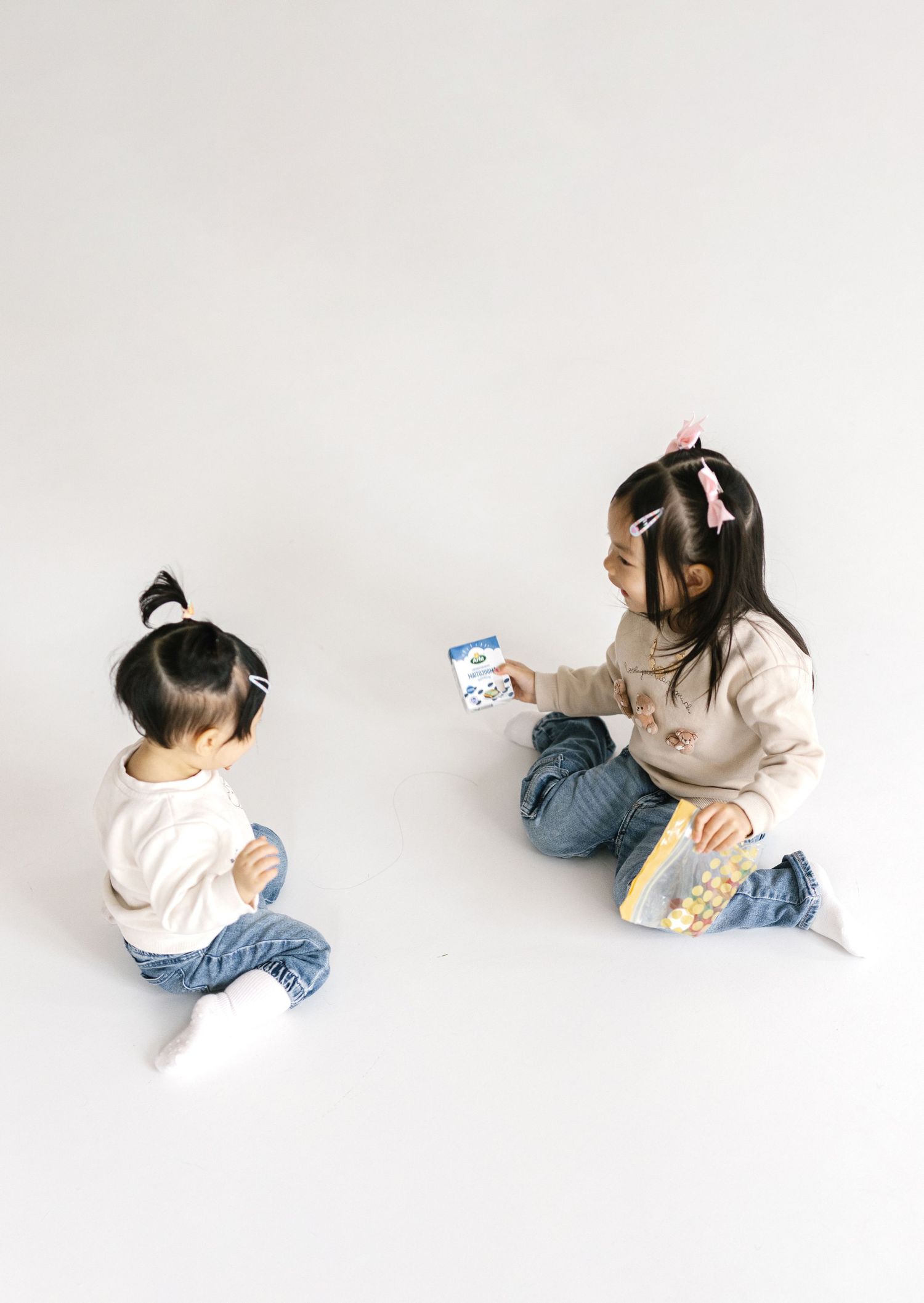 Two young Asian girls sitting on floor, older child showing younger sibling a small book or card.