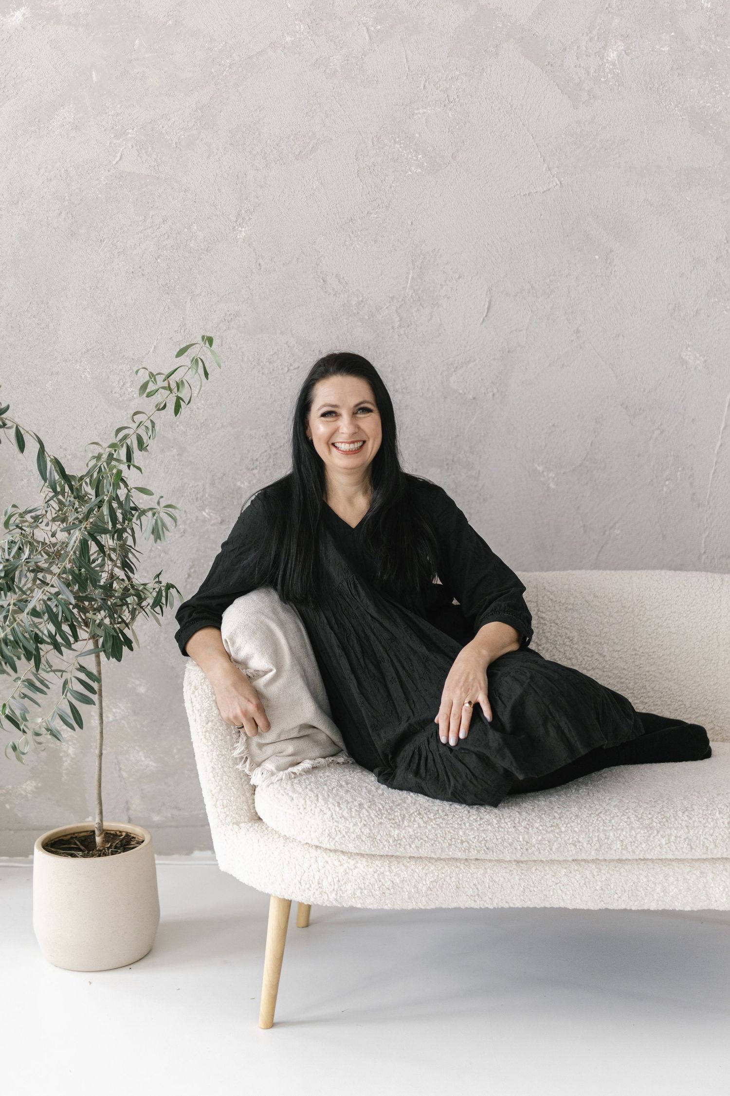 A person in a black outfit reclines on a cream-colored modern chaise lounge next to a potted plant against a gray wall.