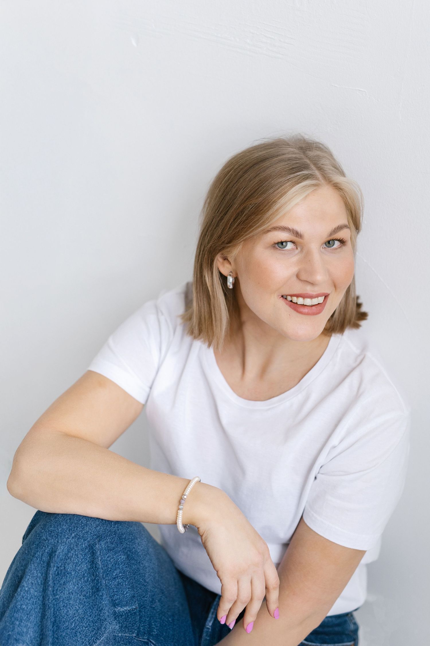 Person in white t-shirt and jeans poses against white wall with natural makeup and jewelry.