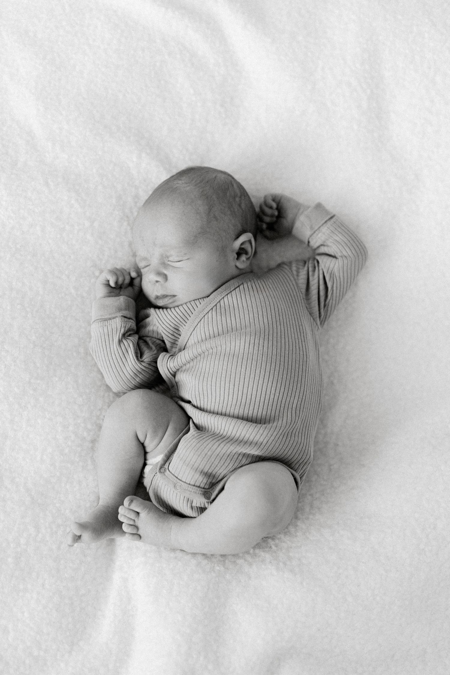 Sleeping newborn baby in ribbed onesie lying peacefully on white blanket in black and white.