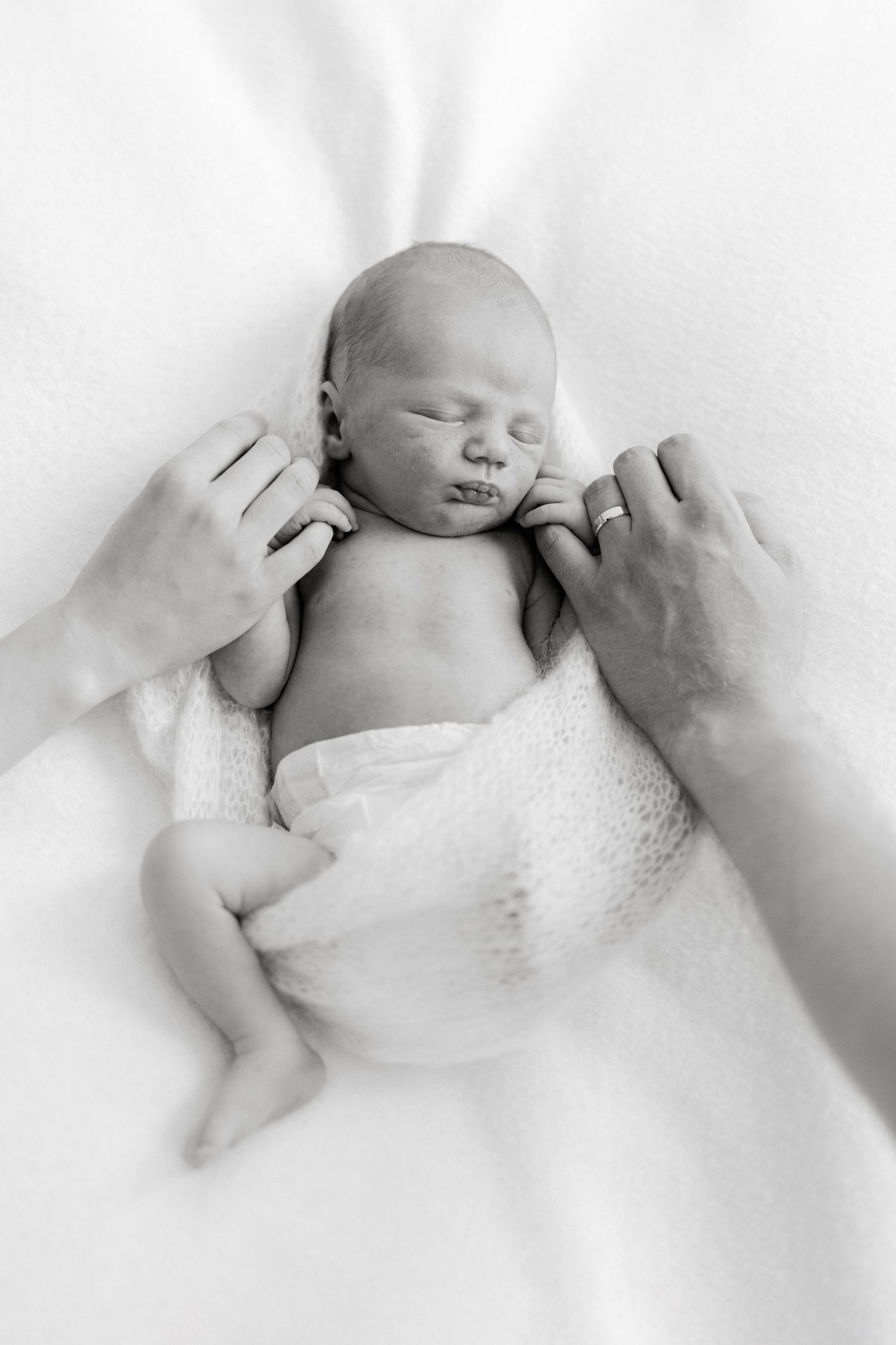 Black and white photo of a sleeping newborn baby wrapped in white, cradled gently by parents hands.