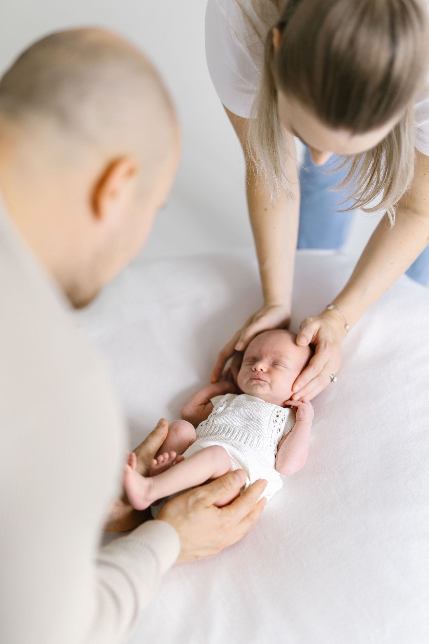 Overhead view of gentle hands cradling a sleeping newborn baby in white clothing.