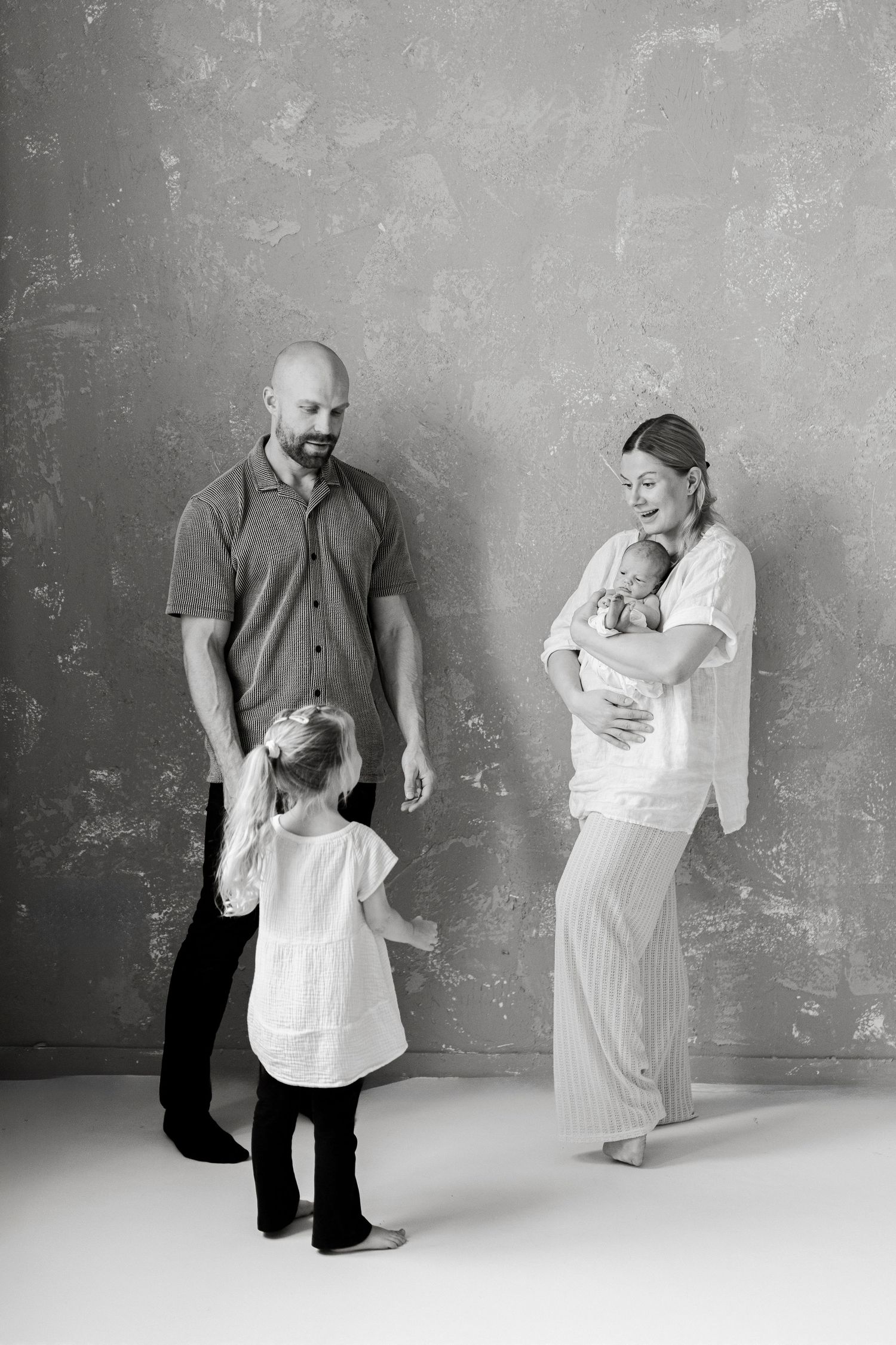 Black and white photo series showing parents cradling a newborn against a textured grey wall.