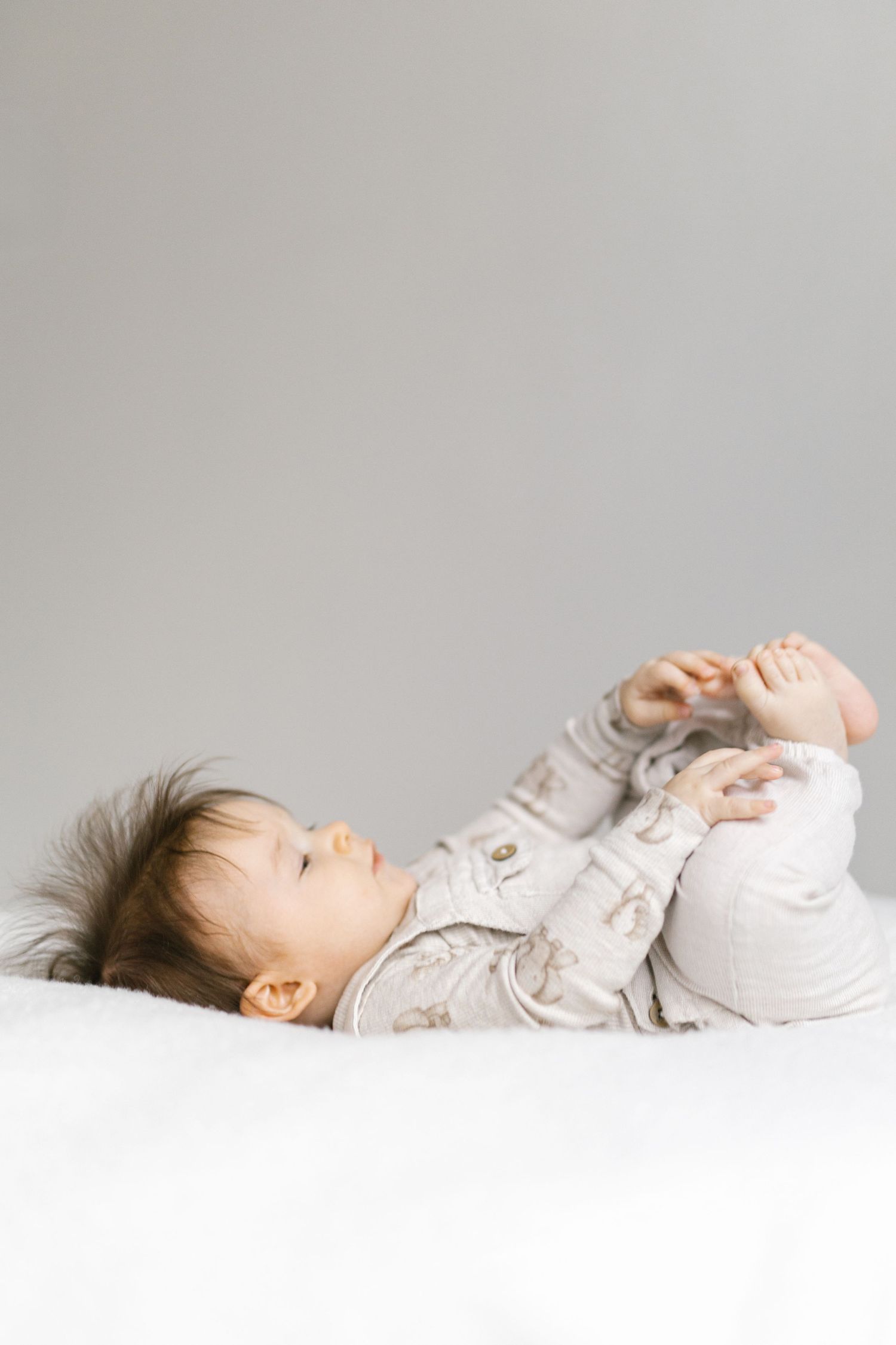 Baby in light colored outfit lying on white surface exploring feet and toes.