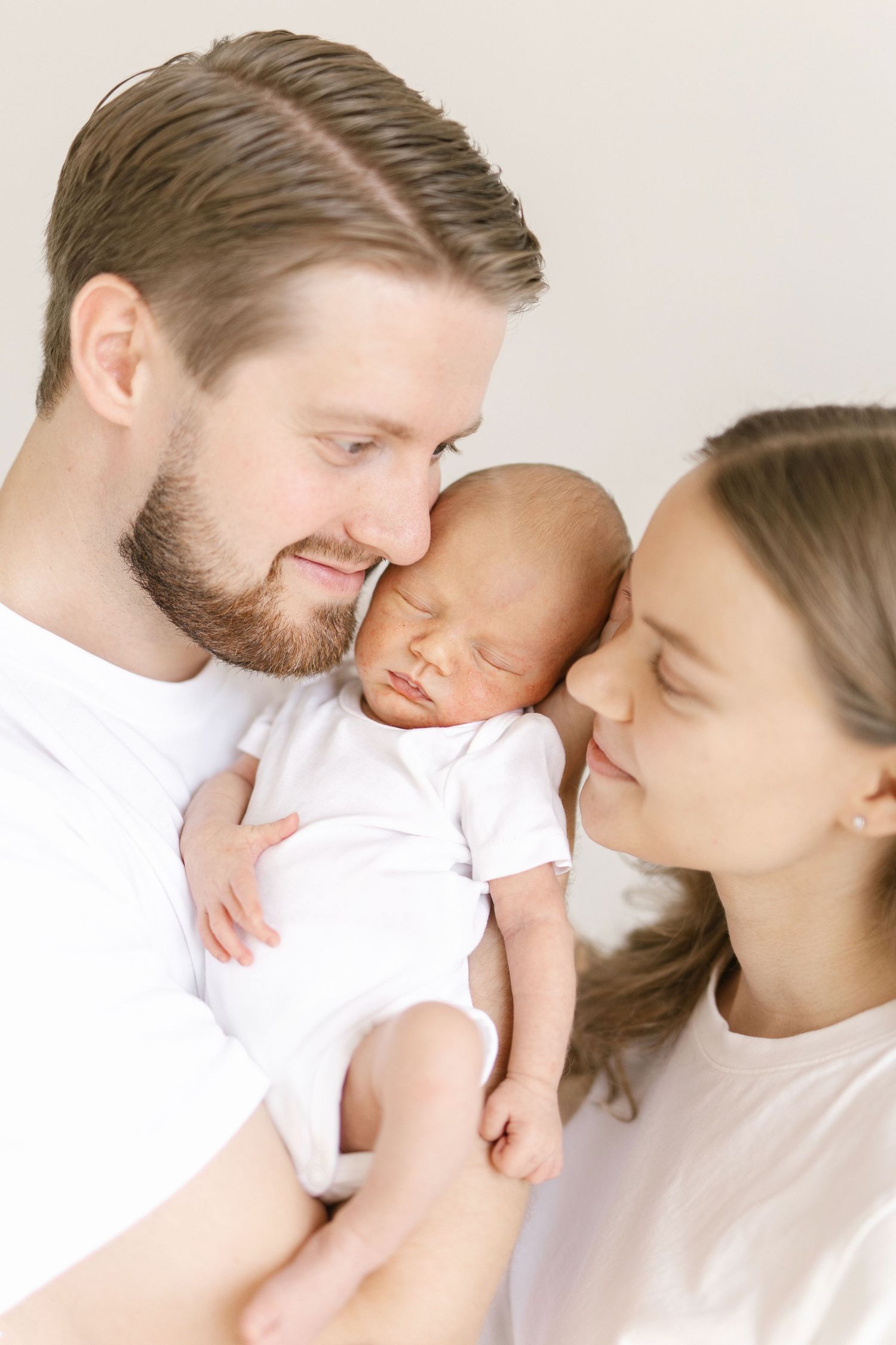 Happy parents holding their sleeping newborn baby, wearing white outfits in a bright, soft-lit family portrait.