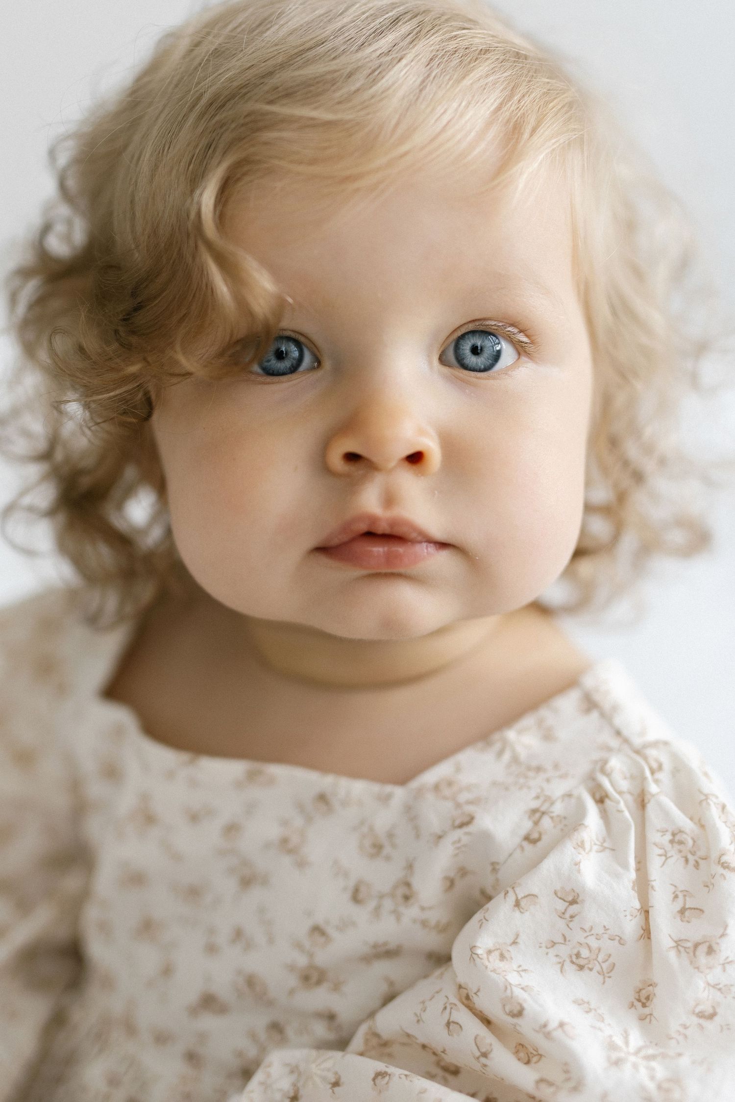 Adorable baby girl with curly blonde hair and blue eyes wearing a white floral dress, portrait photo.