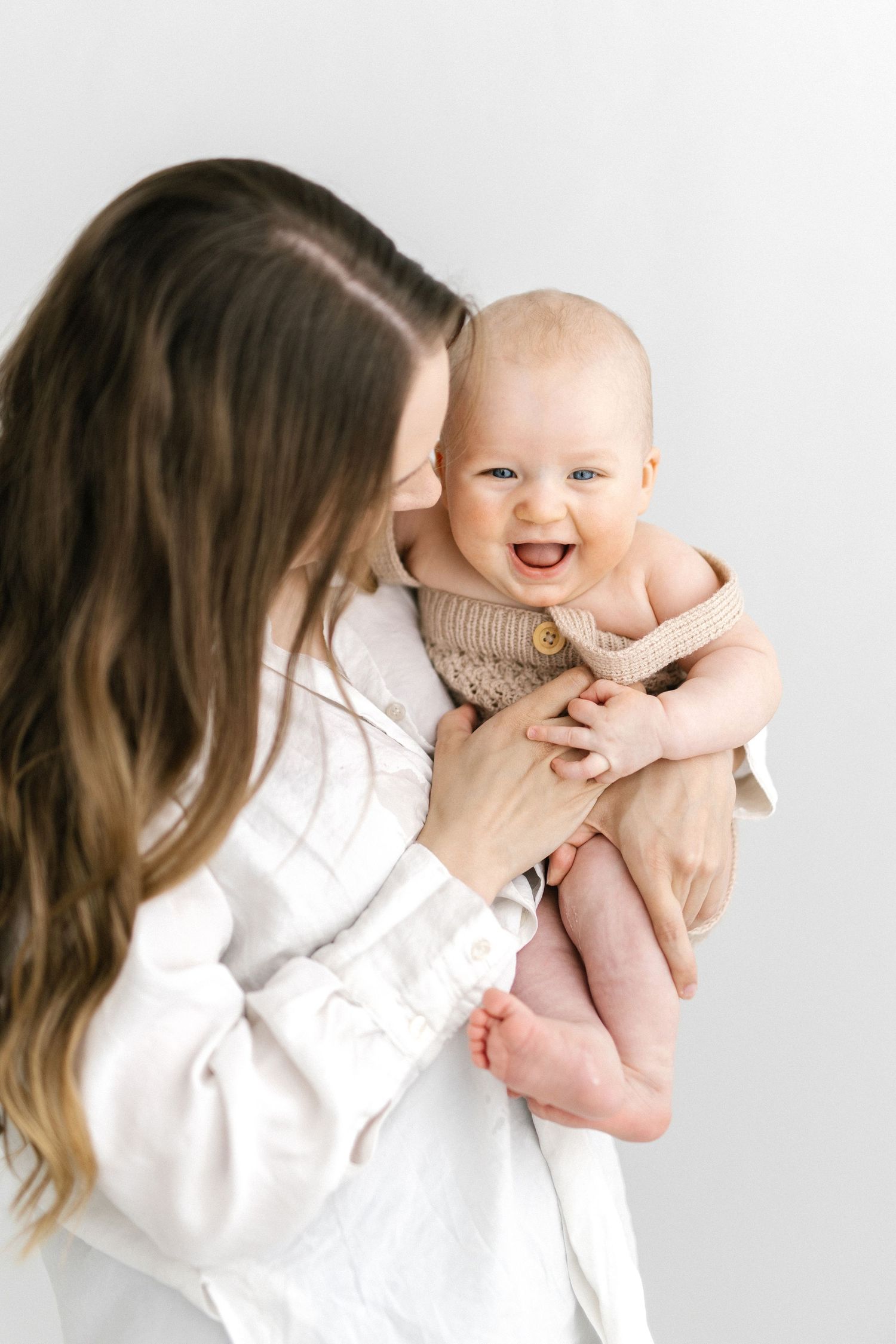 Mother in white top holds laughing baby in knit outfit against a clean white background.